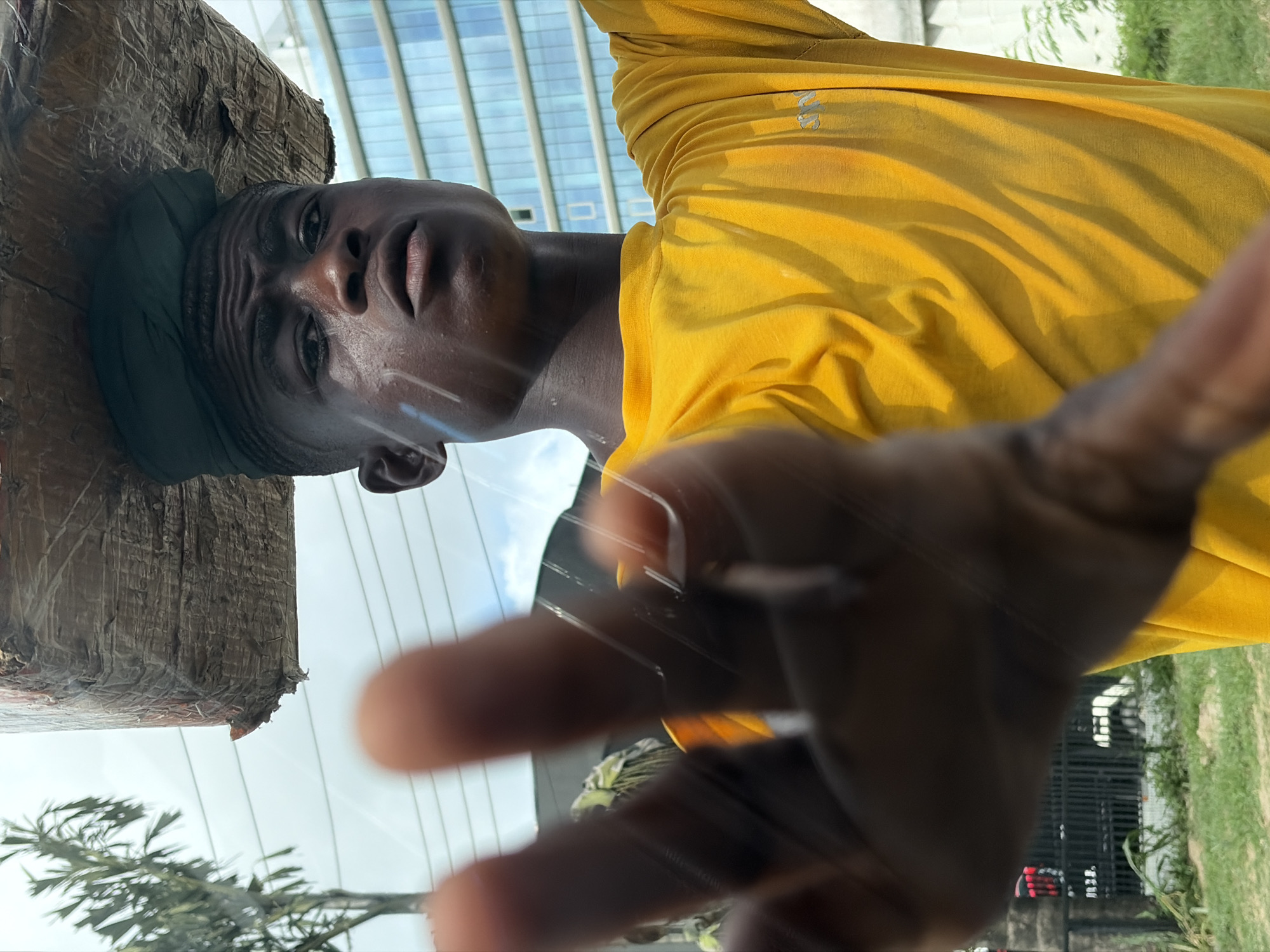 A low-angle view of a man in a yellow shirt and head wrap, positioned beside a rough, wooden post. The man's outstretched hand is toward the camera. In the background, there's a clear sky with modern buildings, suggesting an urban location in Ikoyi, Nigeria. The lower portion shows greenery and a partial view of the landscape.