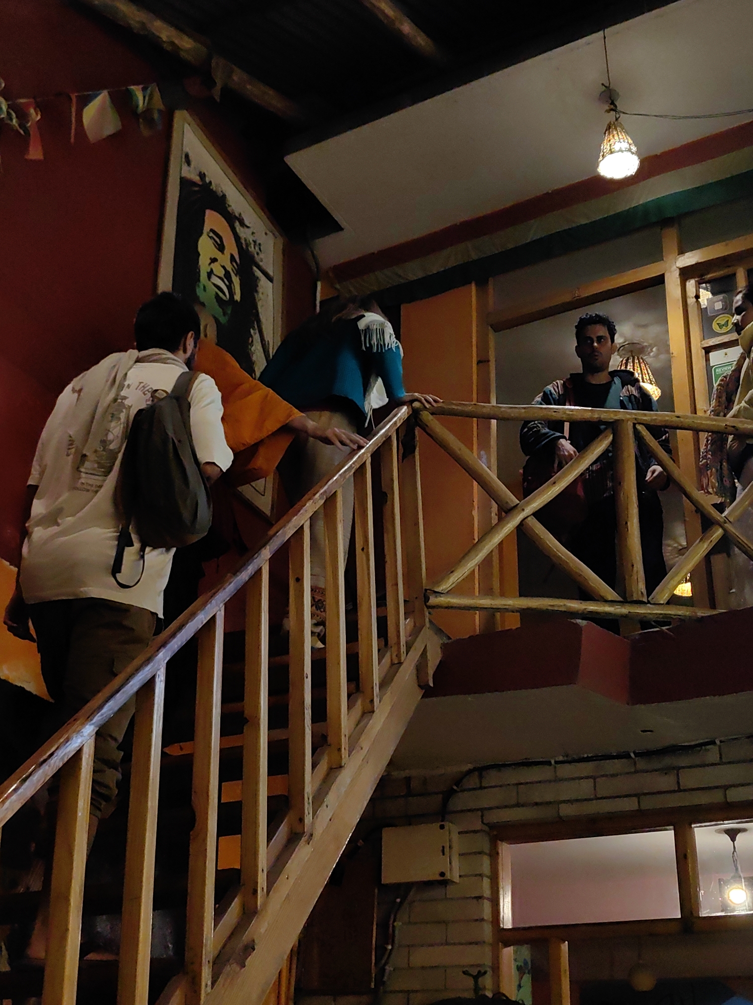 The media file depicts an indoor scene of a dimly lit establishment, possibly a cafe or restaurant, with a rustic wooden interior. In the foreground, a wooden staircase with a slanted railing is prominent. Several people are on and around the stairs.
On the left side of the staircase, a person wearing a white t-shirt and khaki pants is climbing up, carrying a dark backpack. They have a light-colored scarf draped over their shoulders. To their right, another person in an orange garment is also ascending the stairs. Further up the stairs, a person in a teal top and patterned pants is visible.
On a landing above the stairs, a man with dark curly hair is standing, wearing a patterned jacket over a dark shirt and black pants. He appears to be looking towards the viewer. Another person is partially visible to his right, wearing a light-colored garment.
The background features a wall with a large framed painting of Bob Marley. Small triangular flags are strung across the upper part of the wall. A decorative lampshade hangs from the ceiling, casting a warm glow. The walls are made of brick in some sections.
The lighting suggests it is either evening or the interior is intentionally kept dim. The overall atmosphere appears casual and bohemian. No specific text is clearly legible in the image. The presence of the Bob Marley poster and the general aesthetic might suggest a connection to reggae culture or a similar vibe. The location context of Vashisht, India, implies a possible setting in a tourist area or a place frequented by travelers.
