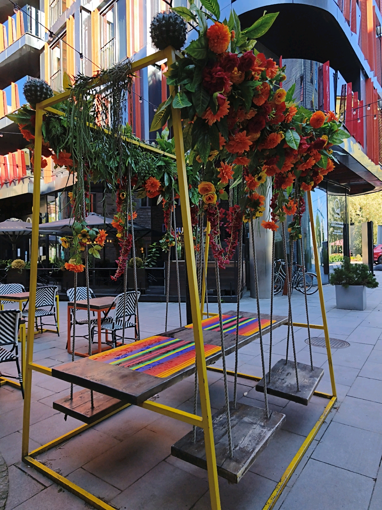 The image shows a yellow swing set with a wooden bench seat painted with colorful stripes, including red, orange, yellow, green, and blue. The swing set is decorated with artificial flowers in shades of orange, red, and yellow, as well as some green foliage and large, dark, spiky orbs that resemble ornamental plants. The swing set is positioned outdoors, on a paved ground made of square gray tiles.
In the background, there are tables and chairs with black and white striped cushions, suggesting an outdoor seating area for a cafe or restaurant. There are also umbrellas providing shade over some of the tables. The building behind the swing set has a modern architectural design with colorful vertical panels in shades of orange, yellow, and red, and a reflective glass facade.
The scene appears to be a public space or an outdoor seating area of a commercial establishment. Based on the architectural style and the context of similar venues, this scene is likely located in the trendy Kalamaja district of Tallinn, Estonia, known for its creative urban spaces and restaurants. No people are visible in the image.