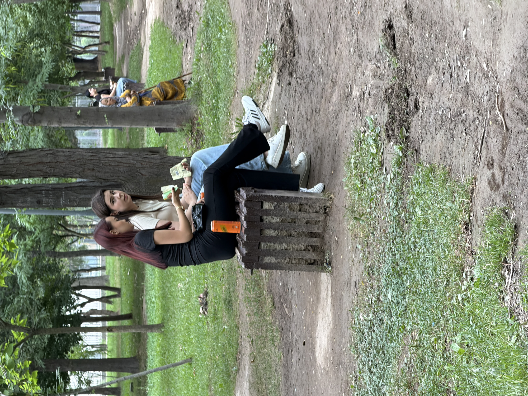 Two young women are seated on a concrete bench in a park. The woman on the left has long, dark red hair and is wearing a black t-shirt and black pants. She is holding some cash in her hands. The woman on the right has long, dark brown hair and is wearing a light-colored t-shirt and blue jeans. She is also holding cash. An orange soda can sits on the edge of the bench. The park has trees and grass, and a path runs through it. In the background, an elderly woman in a yellow and orange garment is seated on another bench, leaning on a cane.