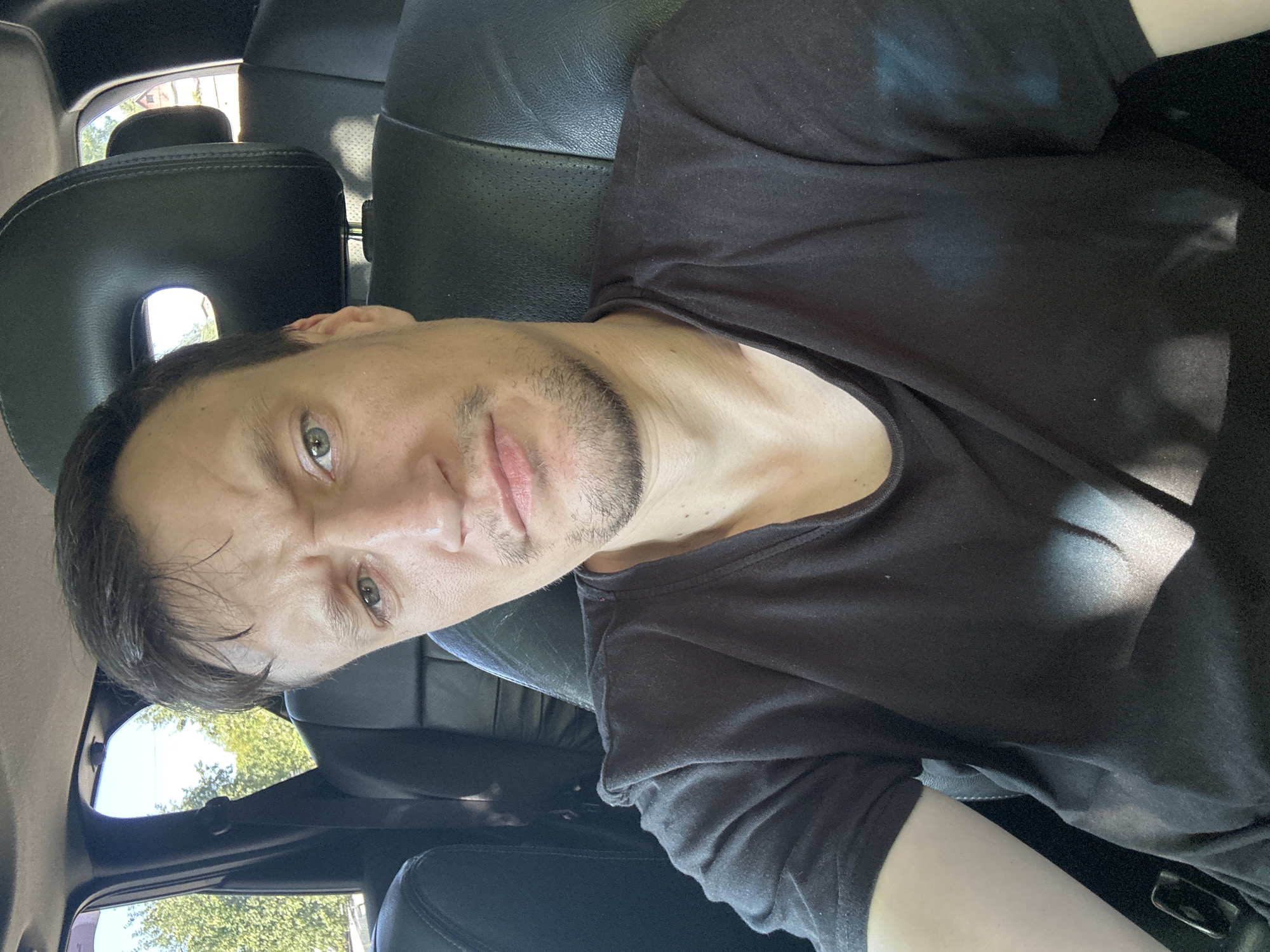 A man with light skin, short brown hair, and stubble is sitting in the driver's seat of a car. He is looking directly at the camera with a neutral expression. He is wearing a black t-shirt. The car interior appears to be black leather. Sunlight streams through the windows, creating shadows on his shirt. The background shows trees and a building, suggesting an outdoor setting, possibly in the city of Makiv, Ukraine.