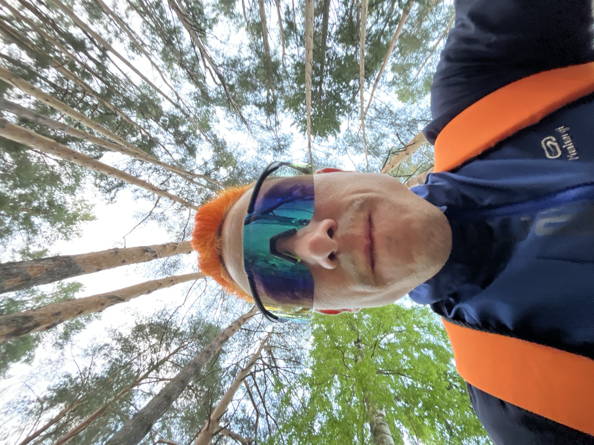 The image is a low-angle selfie of a man, taken outdoors in what appears to be a forest or wooded area near Chaikovsky, Russia. The man has light skin, and orange hair, and is wearing sunglasses. He is also wearing a dark blue jacket with an orange band across the chest/shoulder area. The background consists of numerous tall trees with green foliage and bare branches. The camera is pointed upwards, capturing the tops of the trees against a cloudy sky.