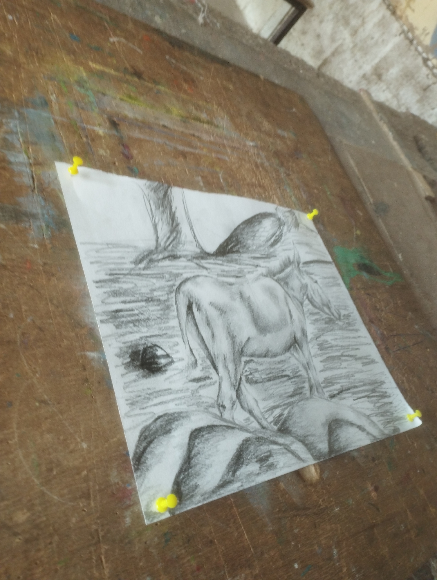The image depicts a charcoal drawing of a semi-nude figure, possibly a woman, hunched over in what appears to be water or a rocky stream. The figure is positioned centrally, with its back to the viewer, showcasing the musculature of the back and shoulders. The drawing is rendered in grayscale with varying degrees of shading to create depth and form. The figure is surrounded by abstract shapes that suggest rocks or a landscape. The drawing is pinned to a wooden surface, likely a workbench or easel, which is weathered and shows remnants of paint splatters in various colors, including blue, yellow, green, and red. The background is out of focus but suggests an indoor setting with a concrete wall and a glimpse of a metal frame. Yellow pushpins are used to secure the drawing to the wooden surface.