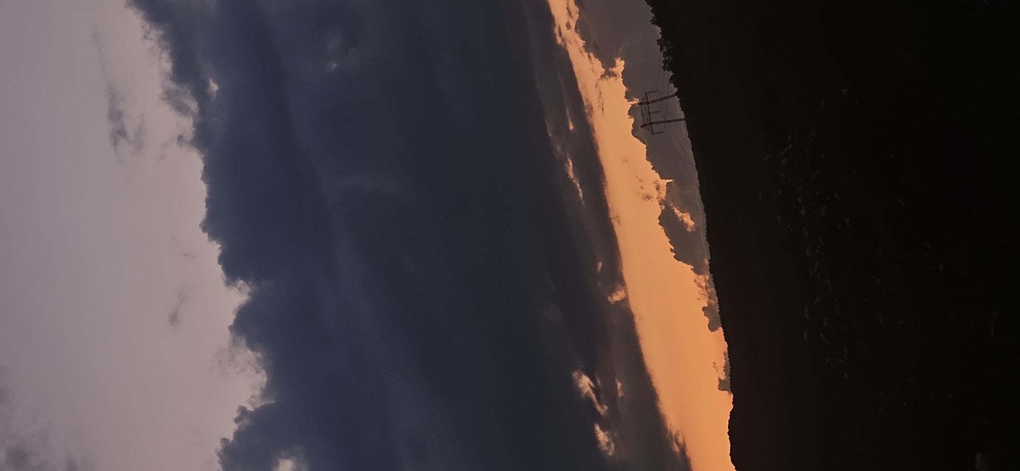 The image displays an outdoor twilight scene in Gutsal, Bulgaria. The sky is bisected by a prominent horizontal band of vibrant orange sunlight, positioned between dark, heavy cumuliform clouds above and a silhouetted landscape below. The clouds in the upper portion are predominantly dark grey-blue, with lighter, purplish hues visible in the uppermost sky. The lower segment of the image reveals a dark, indistinct landmass, likely hills or mountains, covered with dense, dark vegetation, rendered in silhouette against the brighter sky. A single, large electrical transmission tower (pylon) is distinctly silhouetted on the crest of the landmass, positioned on the right side of the frame. No people, animals, or specific actions are present in the depiction.