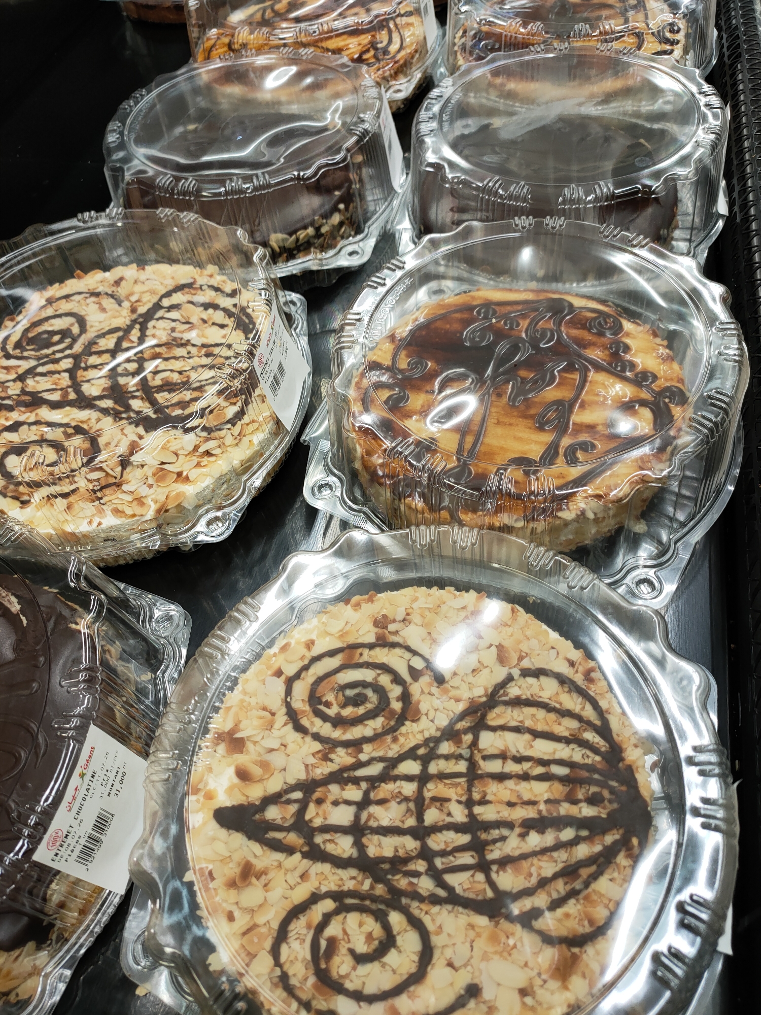 This media file shows an overhead view of several round cakes displayed in clear plastic containers. The cakes are arranged on a dark surface, likely a shelf in a store.
The cakes appear to be of different varieties. One prominently featured cake is covered in sliced almonds and decorated with a dark chocolate drizzle in a swirling pattern. Another cake, partially visible behind it, is also adorned with chocolate icing and what seem to be chocolate shavings or nuts on its sides. Further back, more cakes are visible, some with a striped chocolate topping and others covered in what looks like powdered sugar or a light frosting.
A label is partially visible on the side of one of the cakes, showing text in French and Arabic, including "ENTREMET CHOCOLATINE" and "جيان Geant". The label also includes dates, prices, and barcodes. The lighting suggests an indoor setting, possibly a bakery or a grocery store's dessert section. No people are visible in the frame. The overall impression is a selection of pre-packaged desserts.