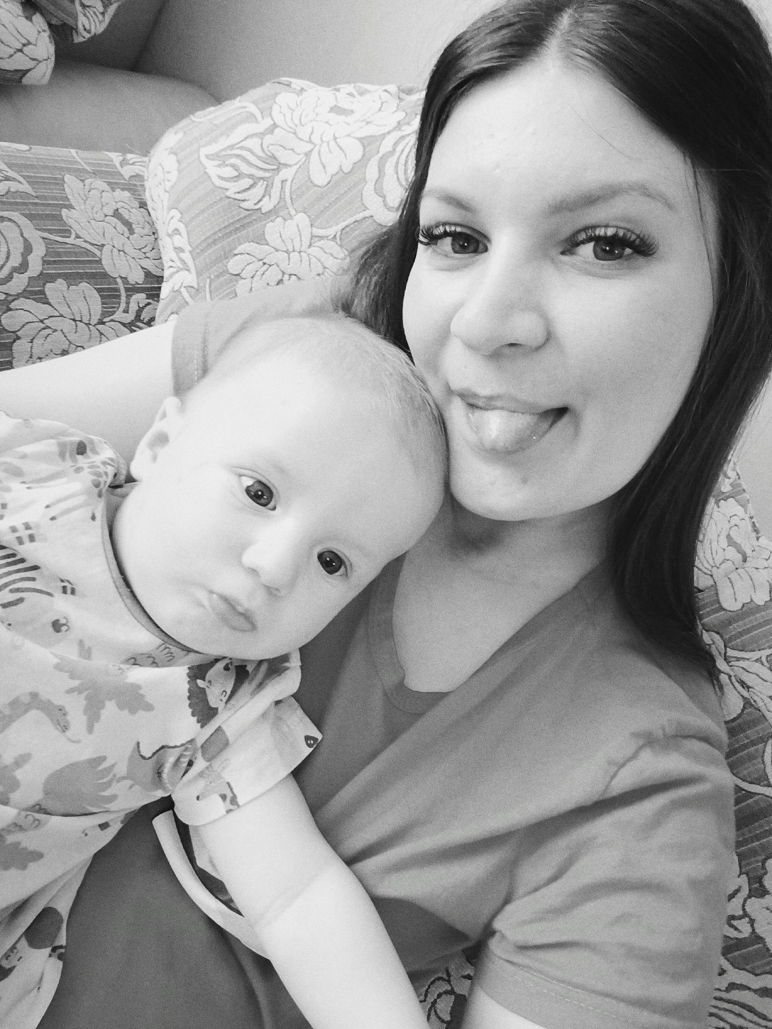 A black and white indoor photograph shows a young woman holding a baby. The woman has long dark hair and is sticking her tongue out slightly. She appears to be wearing false eyelashes and a t-shirt. The baby is wearing a patterned onesie and looking at the camera with a serious expression. They are lying on what appears to be a patterned sofa or bed. No information is present to confirm a location in Tobolsk, Russia.