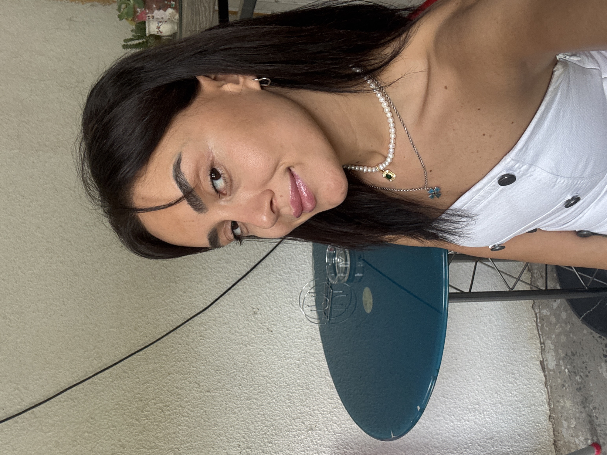 A young woman with dark hair and dark eyes is photographed from the chest up, looking at the camera. She is wearing a white, strapless top with black buttons and two necklaces: one with pearls and a golden charm, and another silver chain with a blue pendant. She has a silver hoop earring in her left ear. Her makeup is subtle, with emphasis on her lips and eyebrows.
Behind her, to her left, is a small, round, teal-colored table with a metal base. On the table, there is a glass ashtray and a metal holder for napkins or cutlery. The background wall is textured and painted a light beige color. A dark, thin wire runs diagonally across the upper left quadrant of the image. To the right of the woman's head, a shelf with small potted plants is visible. The floor appears to be concrete.
The scene appears to be outdoors, possibly on a balcony or patio. The lighting suggests it is daytime. There is no indication of the specific location within the city of Tbilisi, Georgia.