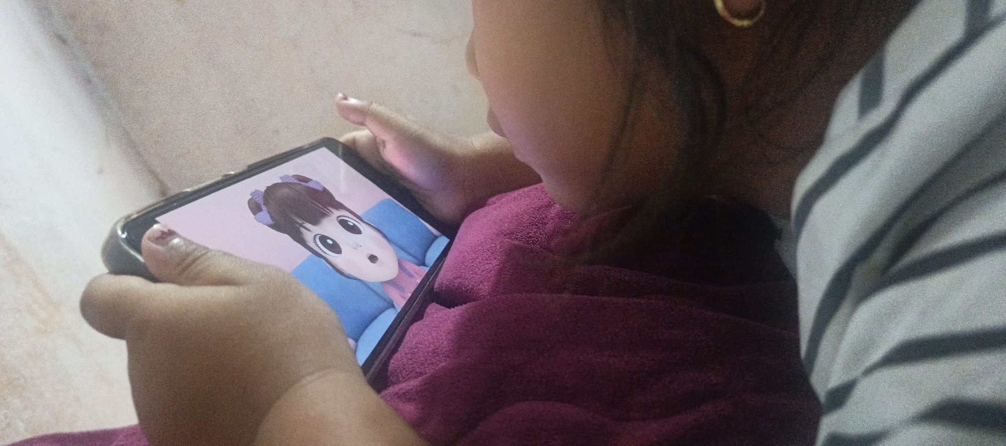 A child, viewed from the side and over the shoulder, holds a smartphone displaying a cartoon character with large eyes and a surprised expression. The child is wearing a dark pink garment and their fingers are visible holding the device. In the background, a striped garment and what appears to be a wall are visible. The scene is indoors, likely in a residential setting in Cinta Dame, Indonesia.