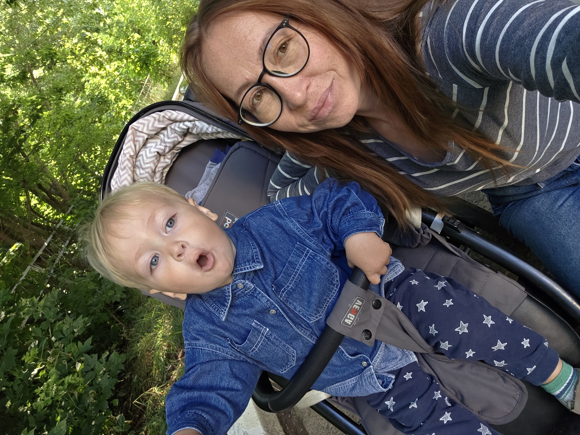 A selfie taken from a slightly high angle shows a woman with auburn hair and glasses, and a young child with blond hair and blue eyes. The child is seated in a stroller, wearing a denim shirt and navy pants with white stars. The woman is wearing a striped long-sleeved shirt. The background consists of green trees and foliage, suggesting an outdoor setting. The woman is smiling at the camera, and the child has an open-mouthed expression. The stroller is grey and has a patterned lining. The scene appears to be set in a park or a natural area. There is no specific indication of the city of Chaikovsky, Russia, within the image itself.