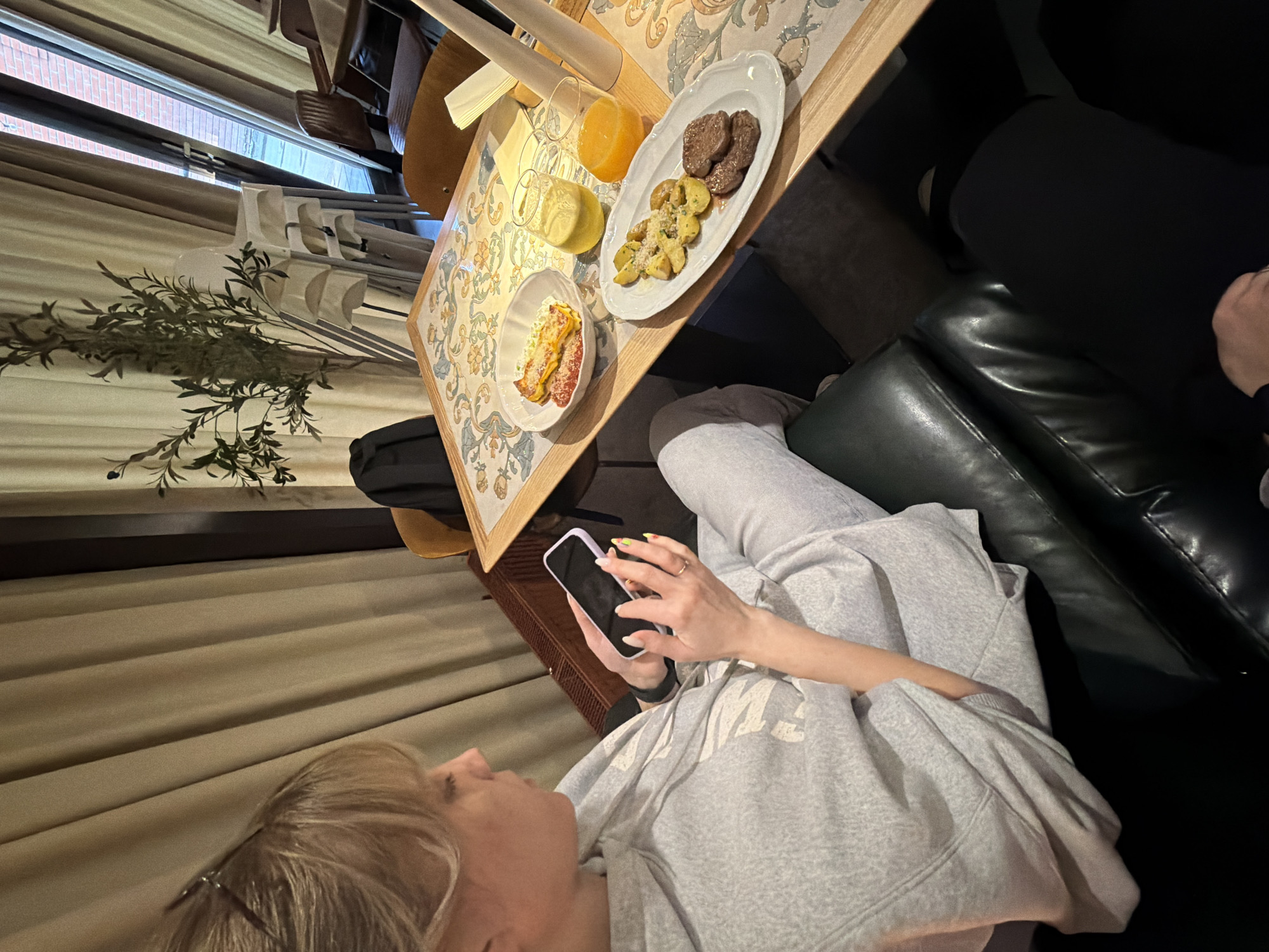 The image depicts an indoor scene within a cafe or restaurant, specifically identified as a Redcup cafe in Perm, Russia. In the foreground, a blonde individual, likely female, is seated in a dark green leather armchair, looking down at and operating a light purple-cased smartphone with both hands. She is wearing a grey athletic tracksuit, a black smartwatch on her left wrist, and a gold ring on her right ring finger; her fingernails display multi-colored polish. The lower body of a second individual, wearing black trousers, is partially visible in the bottom right corner, indicating another person is seated opposite.
A rectangular wooden table with a distinctive light-colored floral pattern (featuring blue, green, and orange motifs) occupies the mid-ground. Laid out on the table are several dishes and beverages: a white ceramic bowl contains a baked cheesy dish with red sauce and grated garnish; a white oval platter holds roasted potatoes sprinkled with herbs and three dark oval meat patties; and two glasses contain orange/yellow juices. A white napkin holder with paper napkins is also present.
The background features tall, beige floor-to-ceiling curtains. To the right, a large window reveals a reddish-brown brick building exterior. An artificial plant with dark leaves stands beside a stack of five white plastic high chairs. The overall environment suggests a modern, cozy dining establishment with warm, subdued lighting.