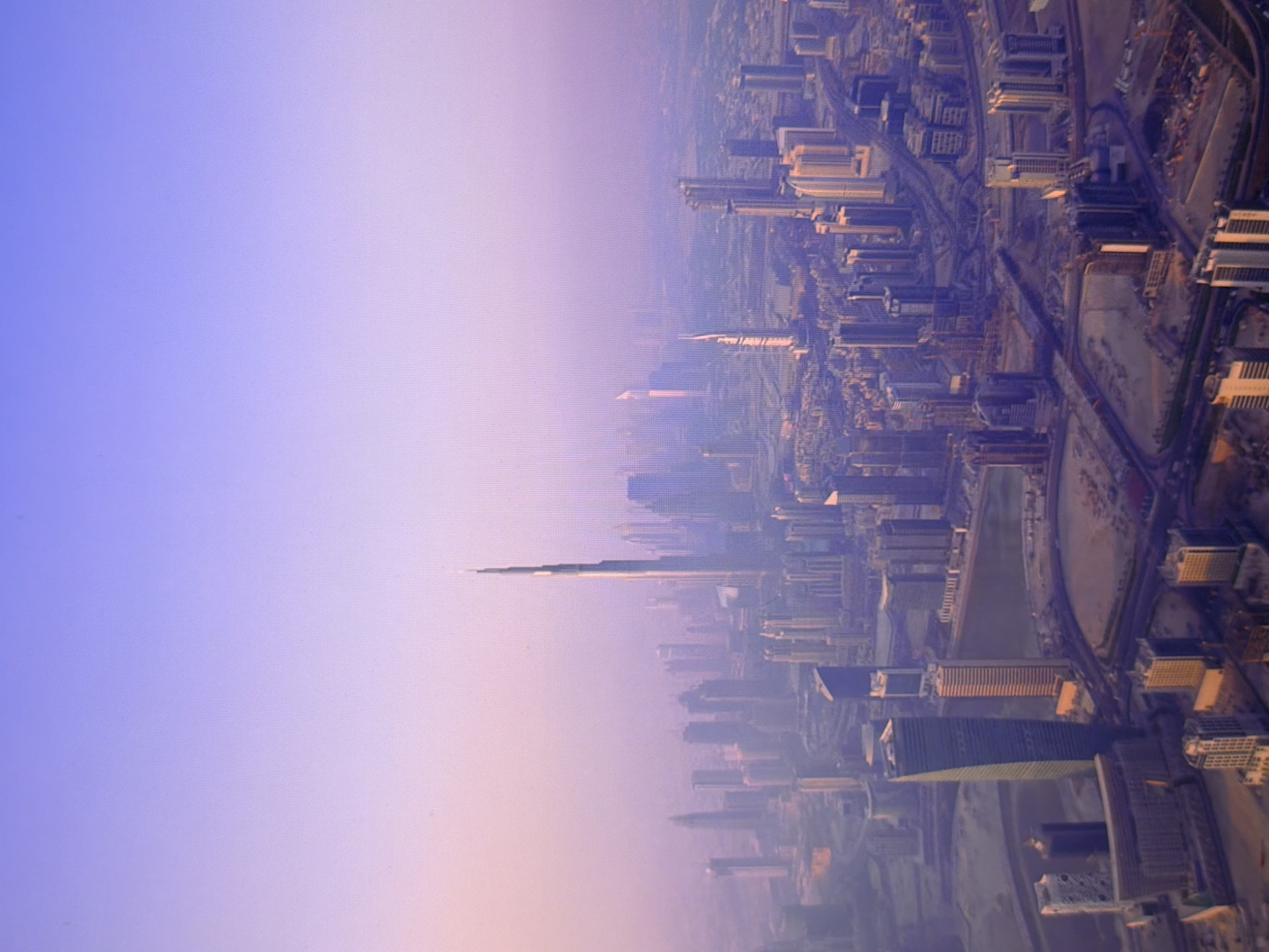 This is a high-angle aerial view of a modern cityscape, likely taken during the late afternoon or early morning due to the soft, hazy light and the purple and pink hues in the sky. The scene is dominated by numerous skyscrapers of varying heights and architectural styles, clustered together and stretching into the distance. In the foreground, several buildings are more clearly defined, with one prominent, tall, and slender skyscraper with a distinctive tapering design, possibly the Burj Khalifa, visible slightly off-center. The urban environment appears to be under development, with some construction sites and areas of exposed earth visible amongst the buildings. Roadways and infrastructure weave through the city, connecting the various structures. The overall impression is one of a dense, high-rise metropolis with a vast skyline. There are no people or discernible animals visible in this image. The sky transitions from a light purple at the top to a softer pinkish-orange near the horizon. The image does not depict the city of Kyiv, Ukraine.