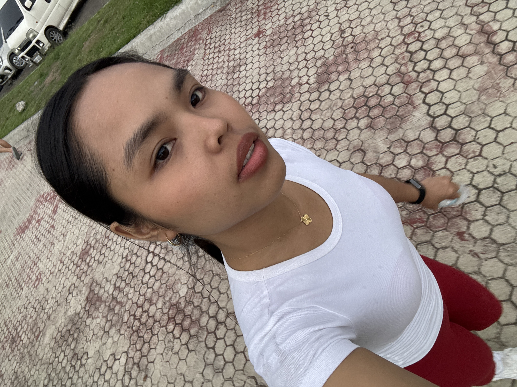 A woman, appearing to be of East or Southeast Asian descent, is the primary subject, captured in a high-angle selfie outdoors in Davao City, Philippines. Her dark hair is pulled back, and she wears a plain white short-sleeved top and red leggings. She accessorizes with a small gold-colored hoop earring, a delicate gold necklace featuring a small flower pendant, and a dark smartwatch on her left wrist. Her expression is neutral, with her gaze directed towards the camera and her lips slightly parted. Faint sweat marks on her white top suggest she may be engaged in or has recently completed physical activity.
The scene is set in an outdoor public space, likely a park or a paved walkway. The ground is distinctive, covered with hexagonal pavers in reddish-brown and lighter beige/grey tones. To the left, a strip of green grass borders the paved area, with two white vans parked behind it. A small portion of another person's foot is partially visible in the far left background. The bright, even lighting indicates it is daytime. No visible text is present in the image.
