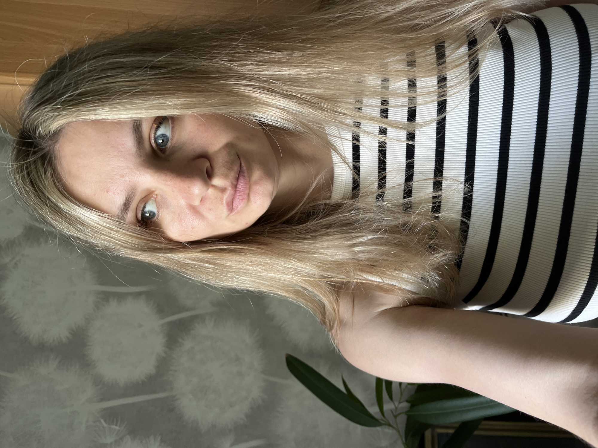 A blonde-haired woman is shown in a close-up selfie. She has light skin and blue eyes and is wearing a black and white striped top. The background includes a wall with a dandelion pattern and a plant with green leaves. The location is not possible to determine, nor is it possible to determine whether the image was taken in Chaikovsky, Russia.