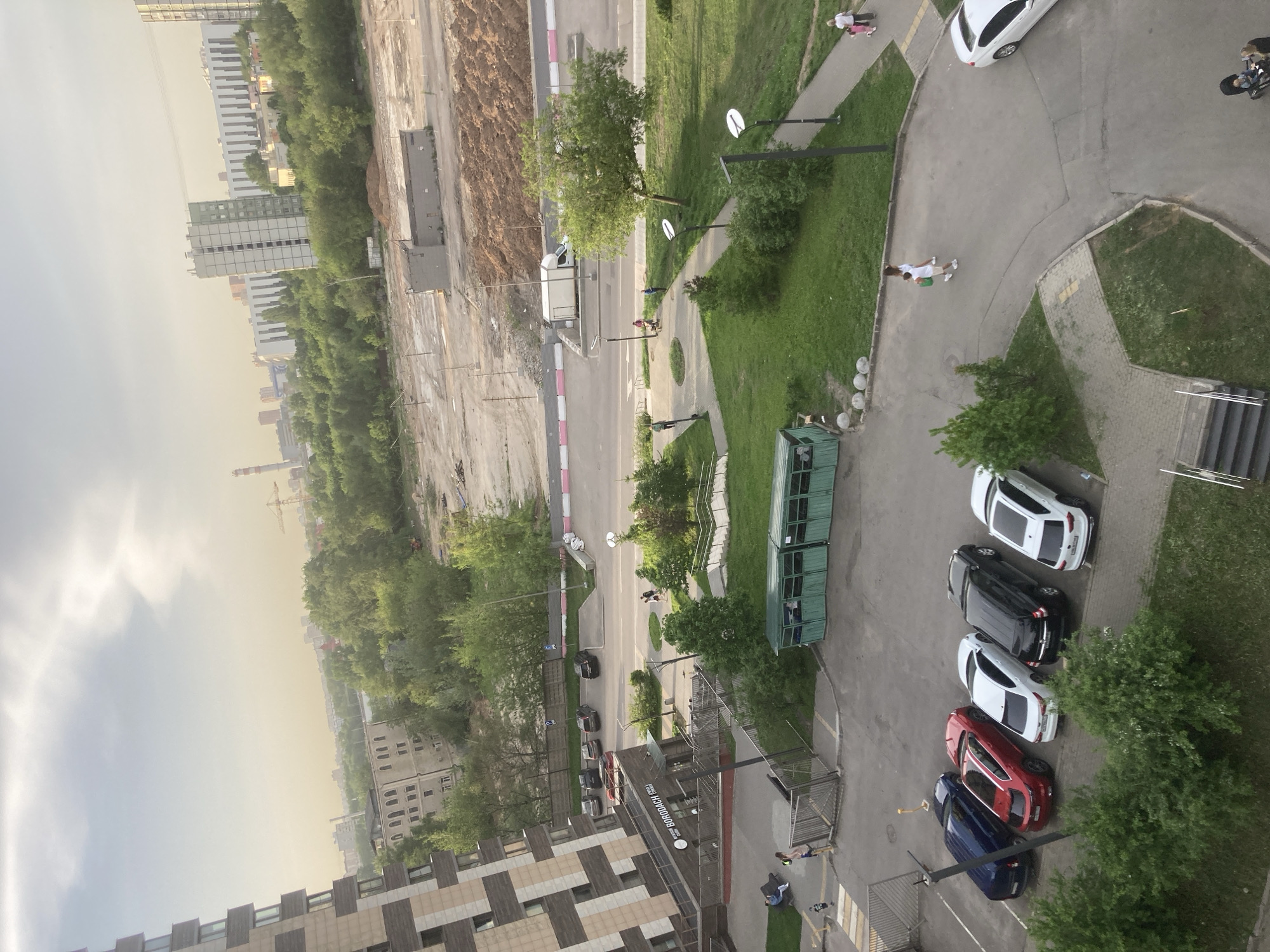 The image, taken from a high vantage point, presents an aerial view of a residential area in Khimki, Russia. In the foreground, a multi-story building with a beige and brown patterned facade is visible. The building appears to have residential units.
Adjacent to the building is a parking area containing several cars parked in a row. The cars are various colors, including blue, red, white, and black. To the right of the parked cars is a patch of green vegetation and trees. A walkway is present.
Beyond the parking area and vegetation, there is a road with pedestrian walkways. Several individuals are visible walking along the sidewalks. There is a construction site in the upper portion of the image, showing exposed soil and construction equipment. The background features other residential buildings and green spaces, blending into the horizon under an overcast sky.