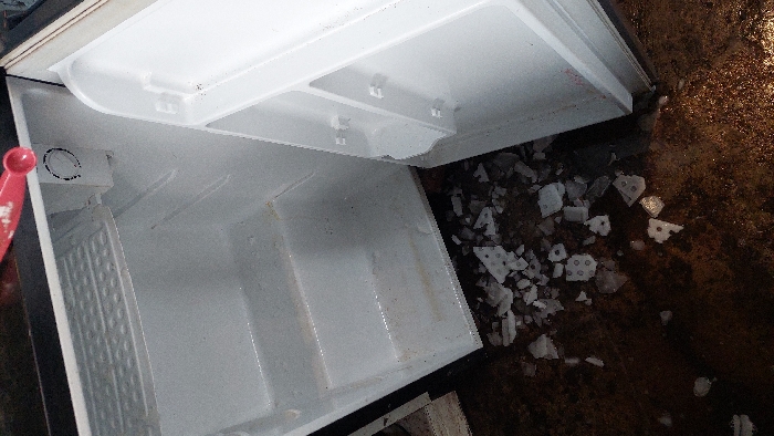 An open, empty compact refrigerator is depicted from an overhead perspective, displaying its white interior with two wire shelves and a circular thermostat control. The interior surfaces show signs of discoloration and moisture. A red plastic spoon rests on the top edge of the refrigerator, partially visible. The dark, wet, and stained floor directly beneath the unit is scattered with numerous broken pieces of white ice and frost, several exhibiting a distinct grid pattern. No individuals are visible. The specific location within Tlaxcala City, Mexico, is not discernible from the visual evidence.