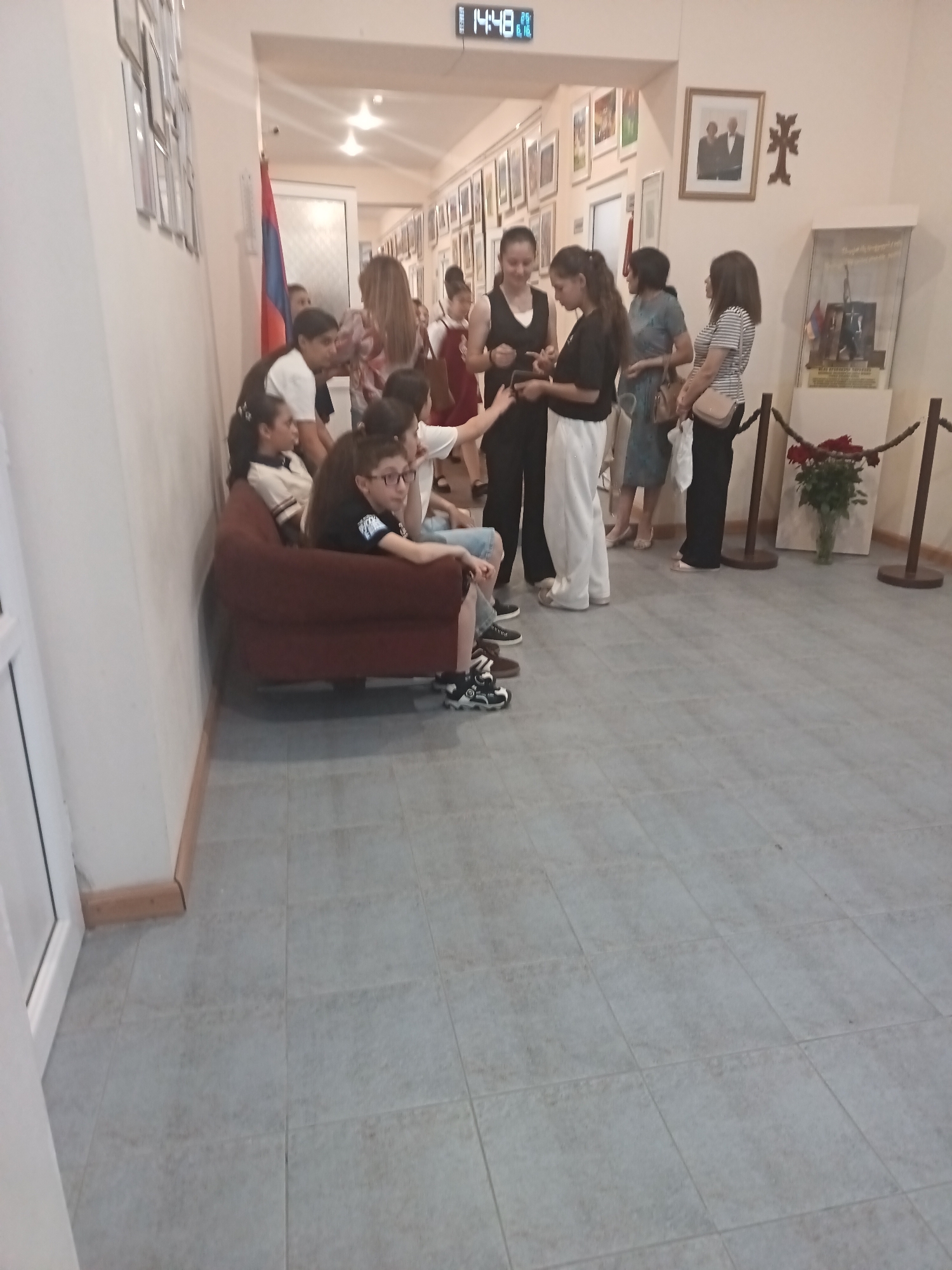 The image shows a group of people, mostly young individuals and some adults, gathered in a hallway or gallery space. The hallway is lined with framed pictures on the walls. There's a digital clock displaying "14:49" at the top of the frame. On the left side of the image, several young people are seated on a dark red sofa. One person is wearing glasses and a black t-shirt, while another is wearing a white and beige striped top. Further down the hallway, more people are standing and talking. An Armenian flag is visible on the left, near a doorway. On the right side of the hallway, there's a display case containing some artifacts, and a bouquet of red roses in a vase. The floor is tiled with light gray squares. The overall atmosphere appears to be that of a public gathering, possibly an event or exhibition.