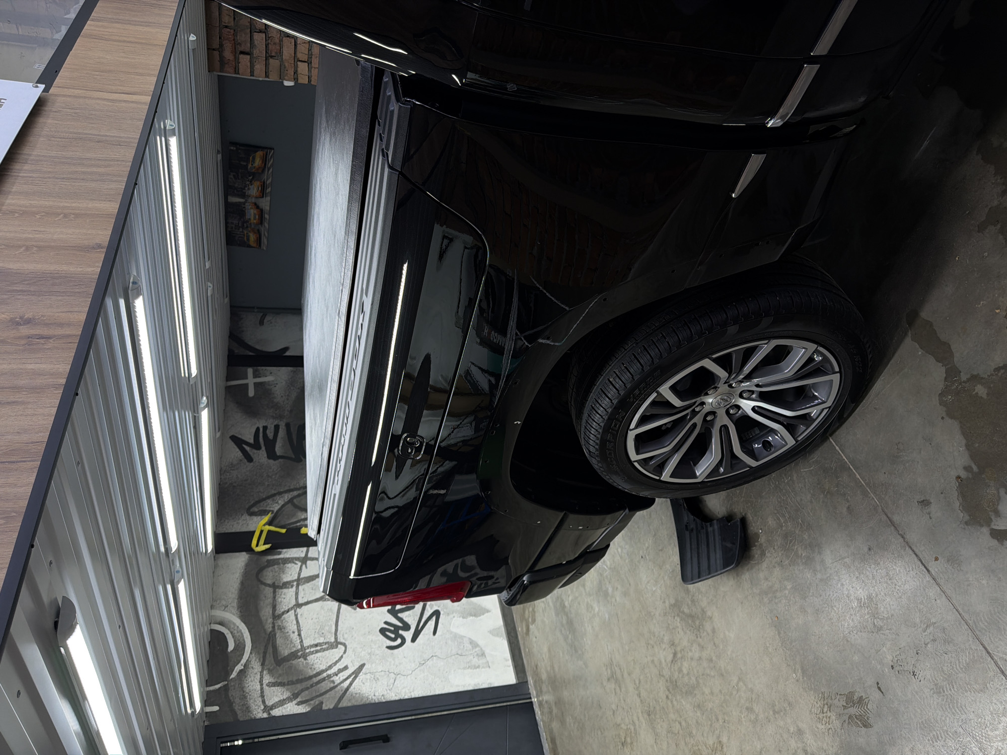 The image shows the rear driver's side of a black pickup truck parked on a concrete floor. The truck has a soft tonneau cover over the bed. The truck's wheel has a multi-spoke rim with silver accents. To the left of the truck is a wall with graffiti art. Above the truck, fluorescent lights illuminate the area. The scene appears to be an indoor garage or workshop. There are no people or visible actions in the image. The location is described as Krasnoyarsk, Russia.