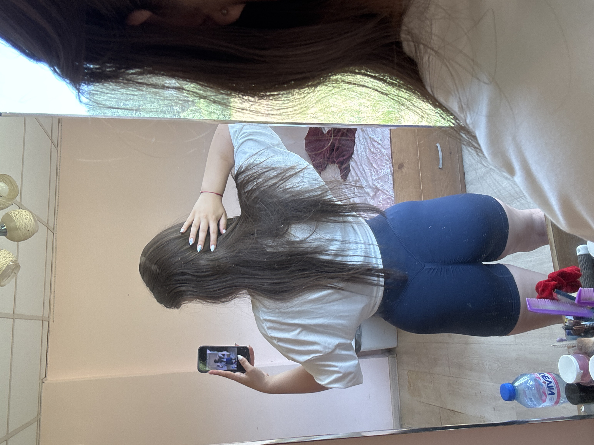 A young woman, seen from behind, is taking a mirror selfie in a room. She has long, dark brown hair and is wearing a white t-shirt and dark blue shorts. She is holding a smartphone in her left hand, which is reflected in the mirror, showing a selfie of a person in a blue top and shorts. Her right hand is touching her hair. In the mirror's reflection, a bed with a patterned comforter and a wooden dresser are visible. A plastic water bottle and various grooming items are on the floor in front of the mirror. The room appears to be residential, with light pink walls and a ceiling with light fixtures. The overall impression is that the photo was taken inside a home. The specific location of Prodanovtsi, Bulgaria, is not determinable from the visual information provided in the image.