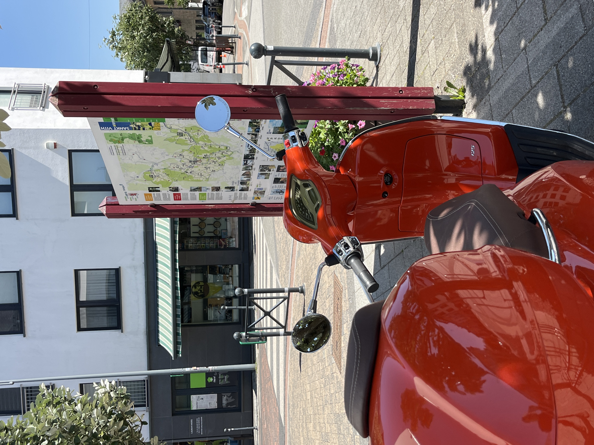 The image depicts a red scooter parked on a cobblestone sidewalk in front of a building with shops. A large map is displayed on a wooden stand, indicating the location is Saint Vith, Belgium. The map shows various routes and points of interest within the region. The scooter is the primary subject, with its reflective surface mirroring the surrounding environment, including trees, buildings, and the sky. The background features typical urban elements such as a crosswalk, streetlights, and other vehicles. The scene is brightly lit by the sun, suggesting it is daytime. There are no people visible in the image.