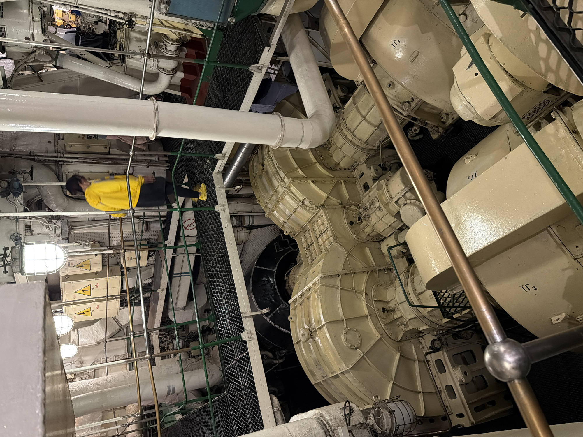 The image depicts the engine room of a ship, possibly a museum ship given the context of the location provided. The room is filled with large, industrial machinery, primarily beige and cream colored, with exposed pipes and electrical components. A person wearing a yellow hooded jacket and black pants is visible on a walkway, looking down at the machinery. Another person's legs are visible further in the background. The environment appears to be indoors, with metal grating walkways and railing systems in place. Lighting fixtures are visible on the ceiling. Based on the provided context, this scene is located in Murmansk, Russia, likely within a ship on display.