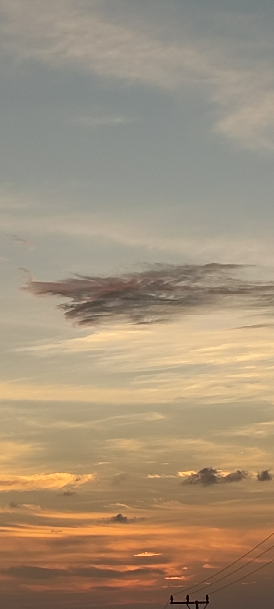 The image depicts a sunset in Lhokseumawe, Indonesia. The sky transitions from a pale blue at the top to a vibrant orange near the horizon. Wispy clouds stretch horizontally across the scene, with a distinct, elongated cloud formation with reddish and gray hues dominating the mid-section. In the lower portion, the orange sky is punctuated by darker, lower-lying clouds. Silhouetted utility poles and wires are visible against the colorful sunset.