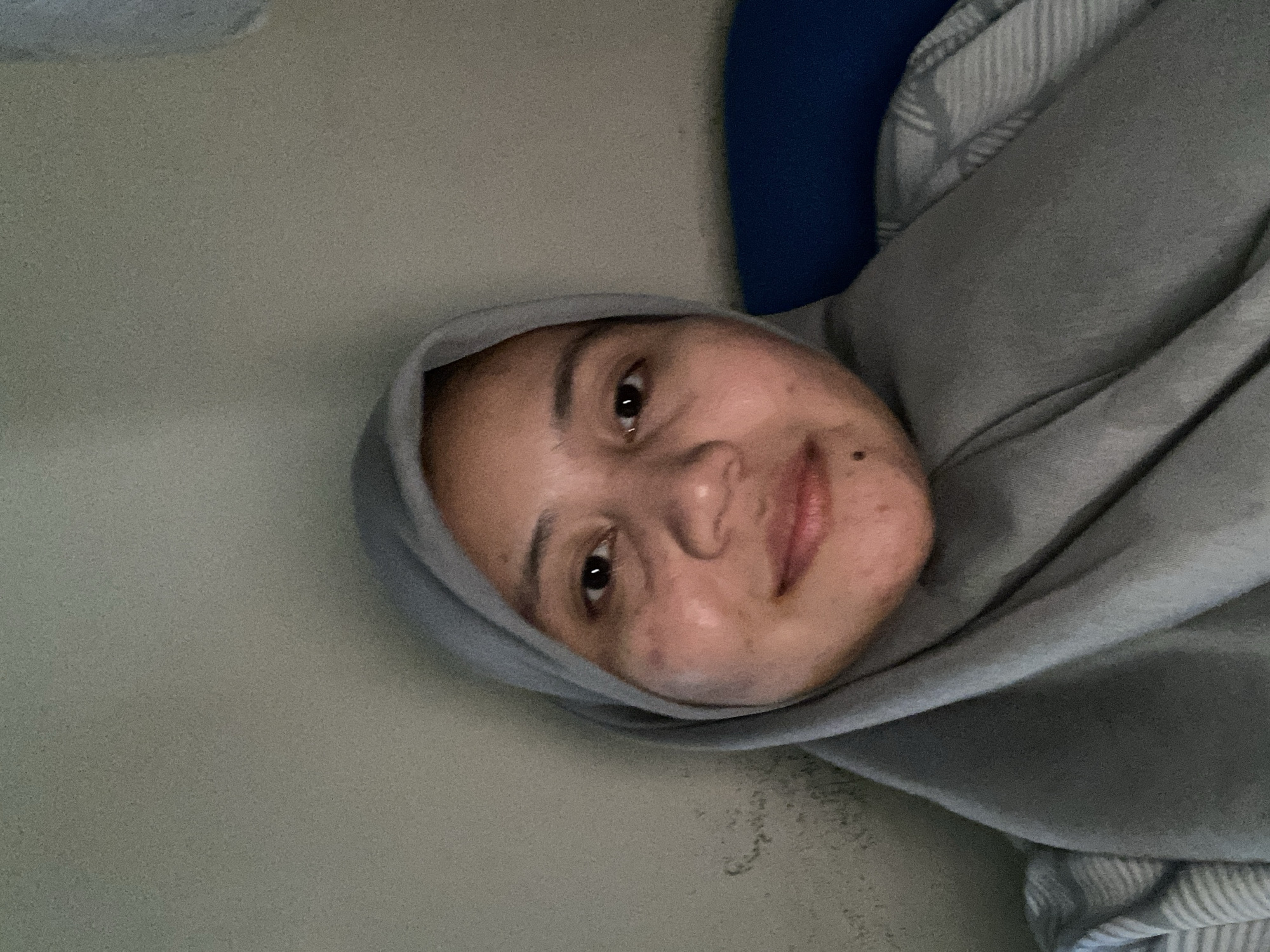 A head and shoulder selfie taken indoors shows a woman wearing a gray hijab. She is looking directly at the camera with her head slightly tilted. Her face is partially illuminated by a light source from the front, highlighting some skin imperfections and light makeup. The background consists of a plain, textured wall and a dark blue surface, possibly a sofa or pillow. The scene does not provide any specific indicators to determine its location within the city of Banjarbaru, Indonesia.