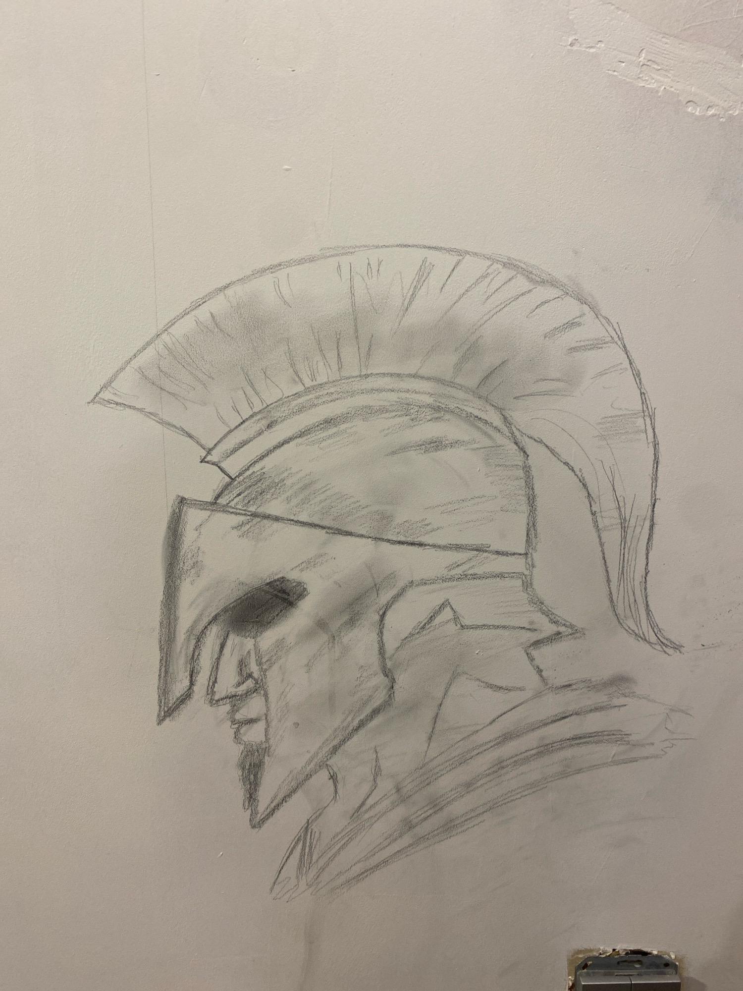 A pencil sketch of a Spartan helmet is drawn on a pale wall. The helmet is depicted in profile, facing left, with a prominent crest of segmented lines extending upwards and backwards. The faceplate features a sharp, angled nose guard and a shadowed eye slit. The lower portion of the helmet extends down, showing layered defenses. The drawing is rendered with varying line weights and shading, suggesting depth and texture. The wall itself appears to be textured, with some slight imperfections visible. There is no indication of Mazlauki, Latvia, or any other specific urban environment associated with this image. The scene is solely focused on the artwork.