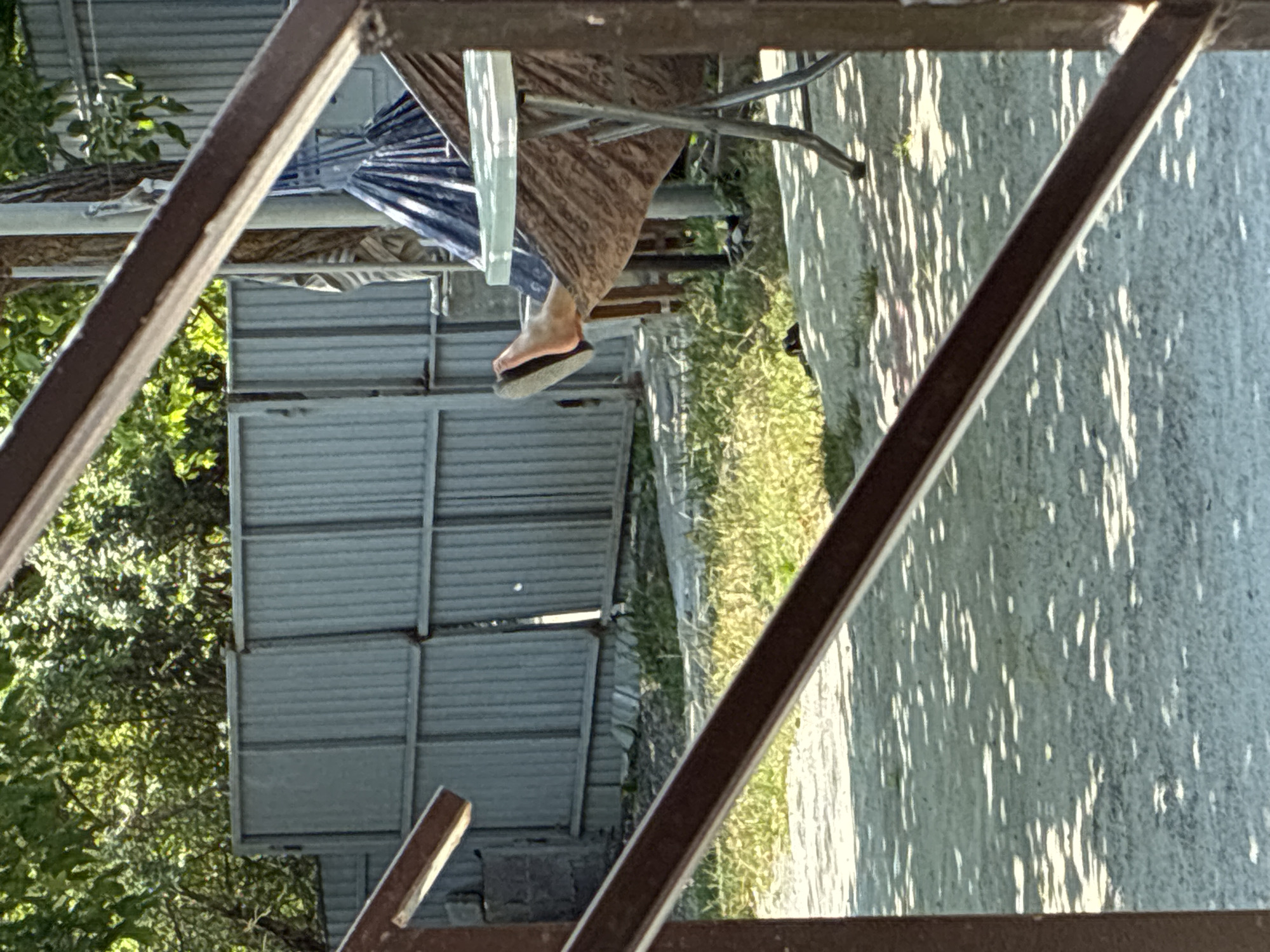 A person is lying in a hammock in the city of Surami, Georgia. The hammock is made of brown fabric with a pattern, and it is supported by a stand. A white-topped table with metal legs is positioned next to the hammock. The person's bare feet are visible, with one foot extending out of the hammock and wearing a flip-flop. Behind the hammock, a corrugated metal fence runs horizontally, partially obscured by trees and foliage. The ground in front of the fence is covered in grass and gravel, with dappled sunlight creating patterns of light and shadow. The foreground is dominated by dark brown, angled railings.