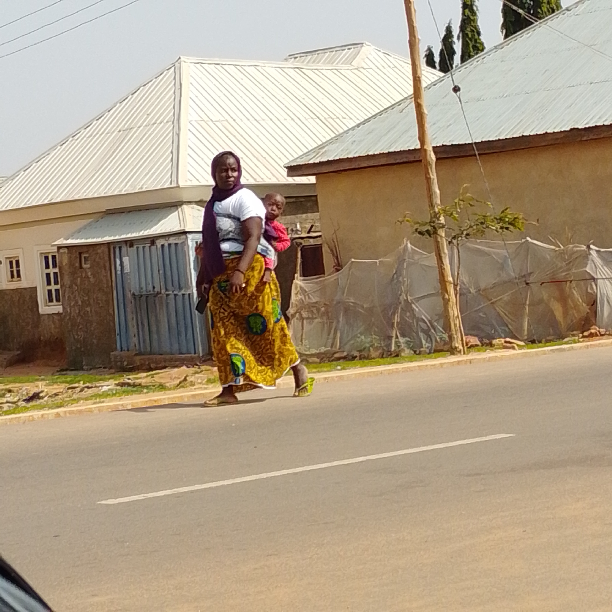 A woman is walking on a paved road in Gombe, Nigeria. She is carrying a young child on her back. The woman is wearing a white t-shirt, a purple headscarf, and a long, yellow and green patterned skirt. She is also wearing flip-flops. The child is wearing a red shirt. In the background, there are buildings with corrugated metal roofs and a tall wooden pole with wires. The road has a white line painted down the center.