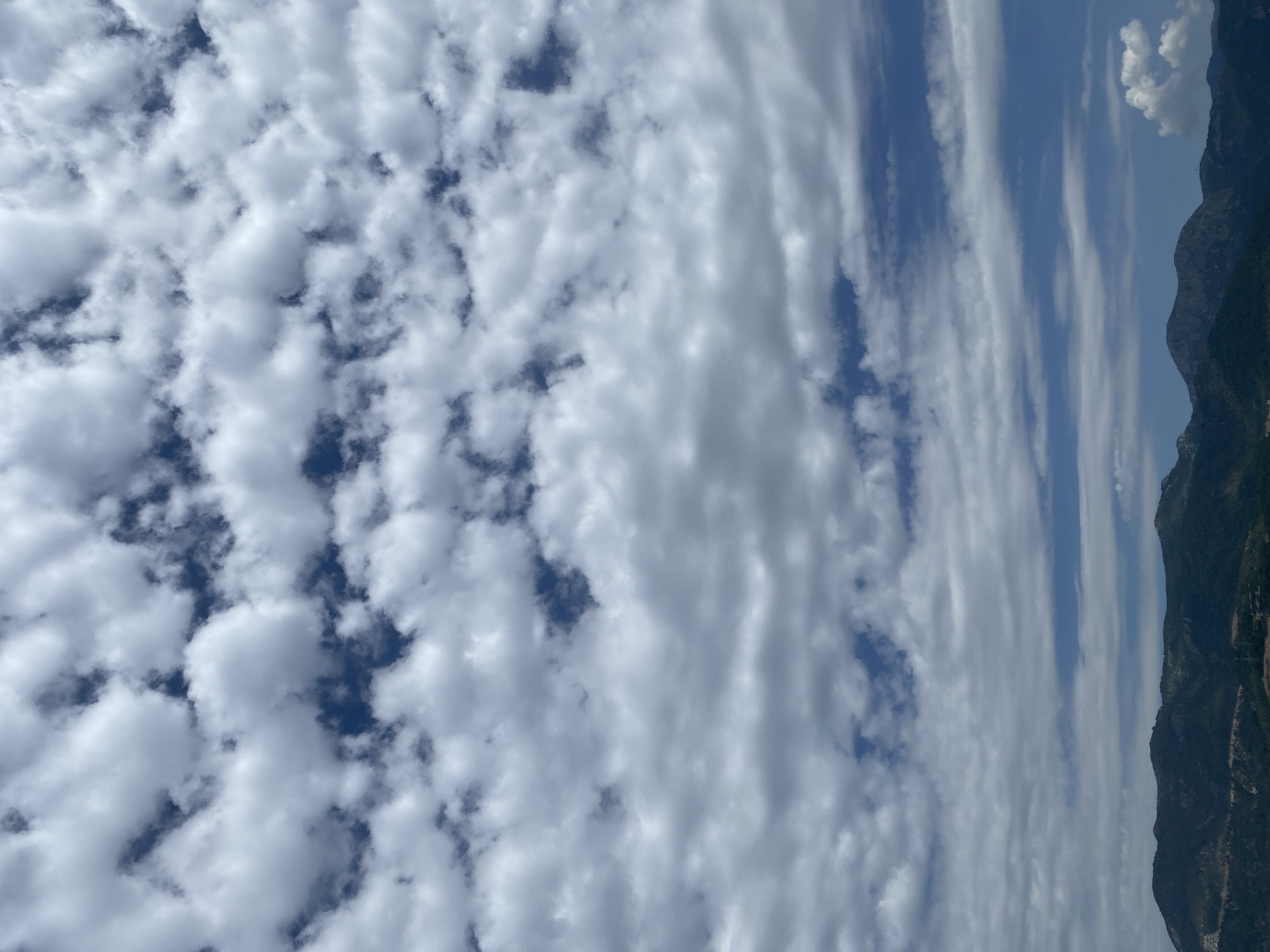 The image is a vertical shot of the sky with mountains on the right side of the frame. The sky is filled with altocumulus clouds with patches of blue visible between them. The mountains appear green and dark. The location is possibly Alanya, Turkey. The time of day seems to be daylight. There are no people visible in the image.