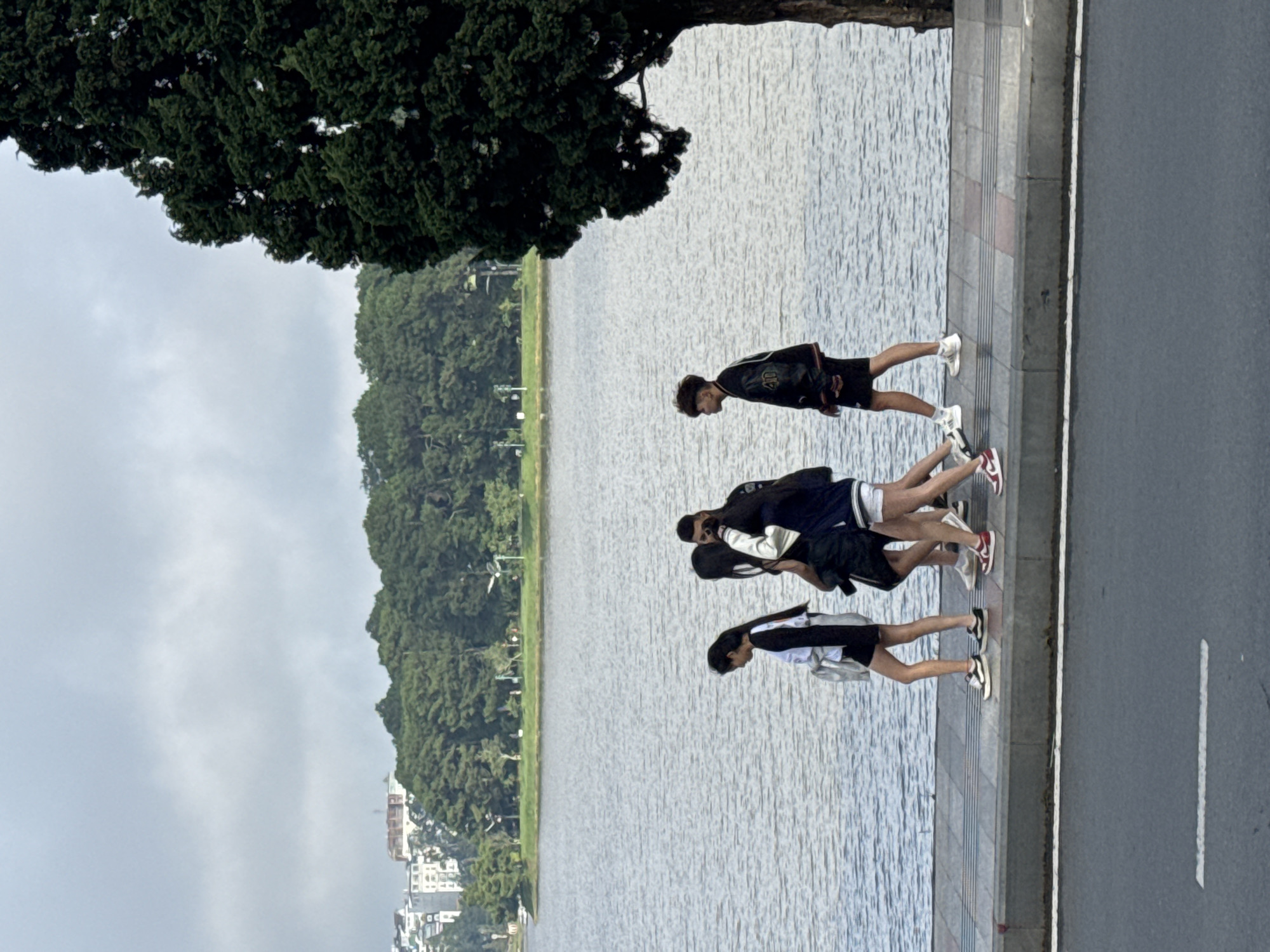 Four individuals, three females and one male, are walking on a paved sidewalk next to a large body of water, likely Xuan Huong Lake in Da Lat, Vietnam. The individuals are walking from left to right, with the male on the far right and the females to his left. They are dressed in casual clothing. The environment consists of the lake, the sidewalk, a tree on the right side of the frame, and a treeline with buildings in the background. The sky is overcast.