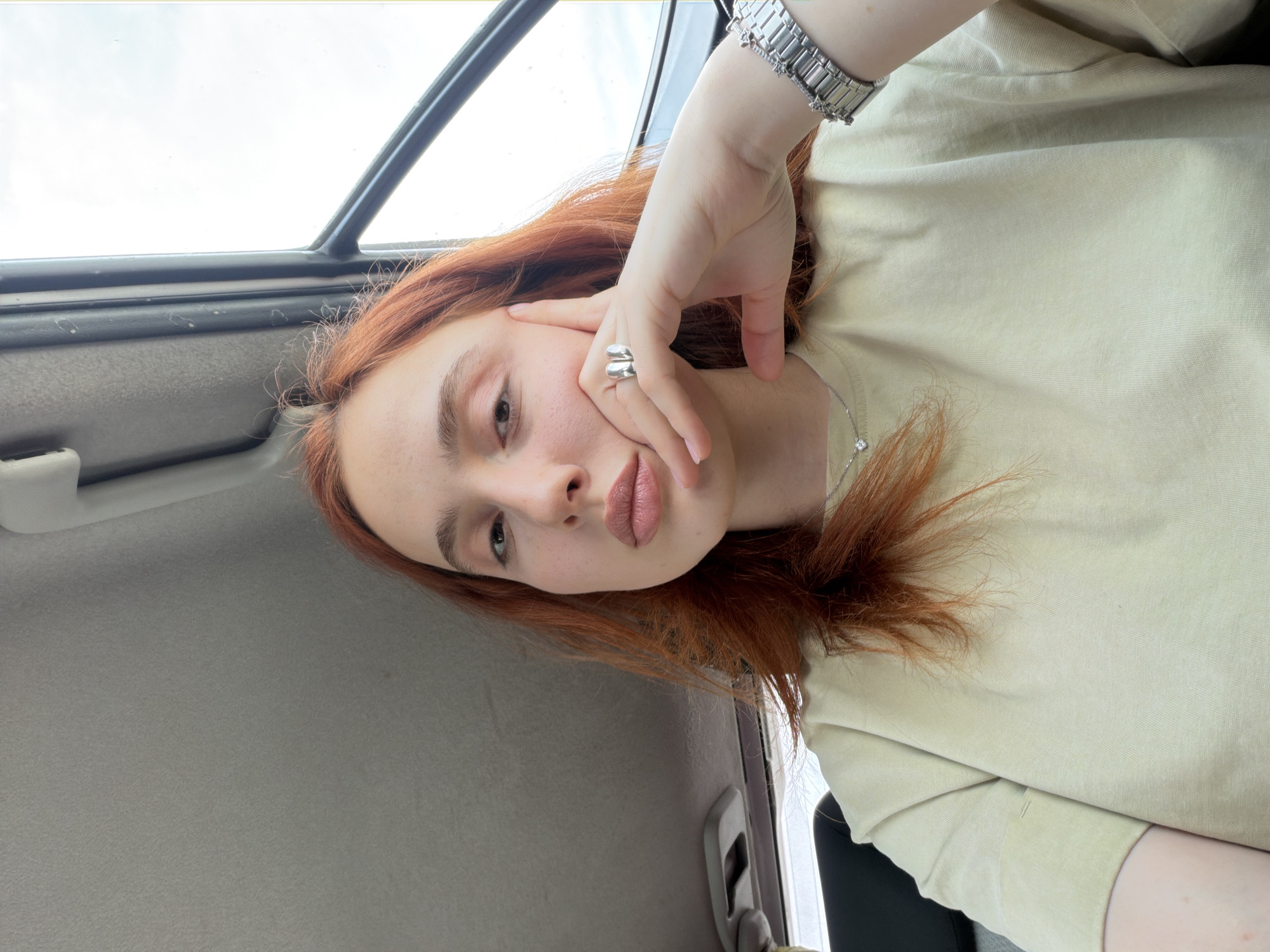 A fair-skinned woman with reddish-brown hair is posing in what appears to be the interior of a car. She is wearing a pale-yellow t-shirt, a silver wristwatch, and a large silver ring on her right hand. Her right hand is positioned near her face, with fingers slightly touching her cheek. The car interior includes a gray door panel and a window with a partial view of the sky. There is no other information suggesting the location in the city of Tbilisi, Georgia.
