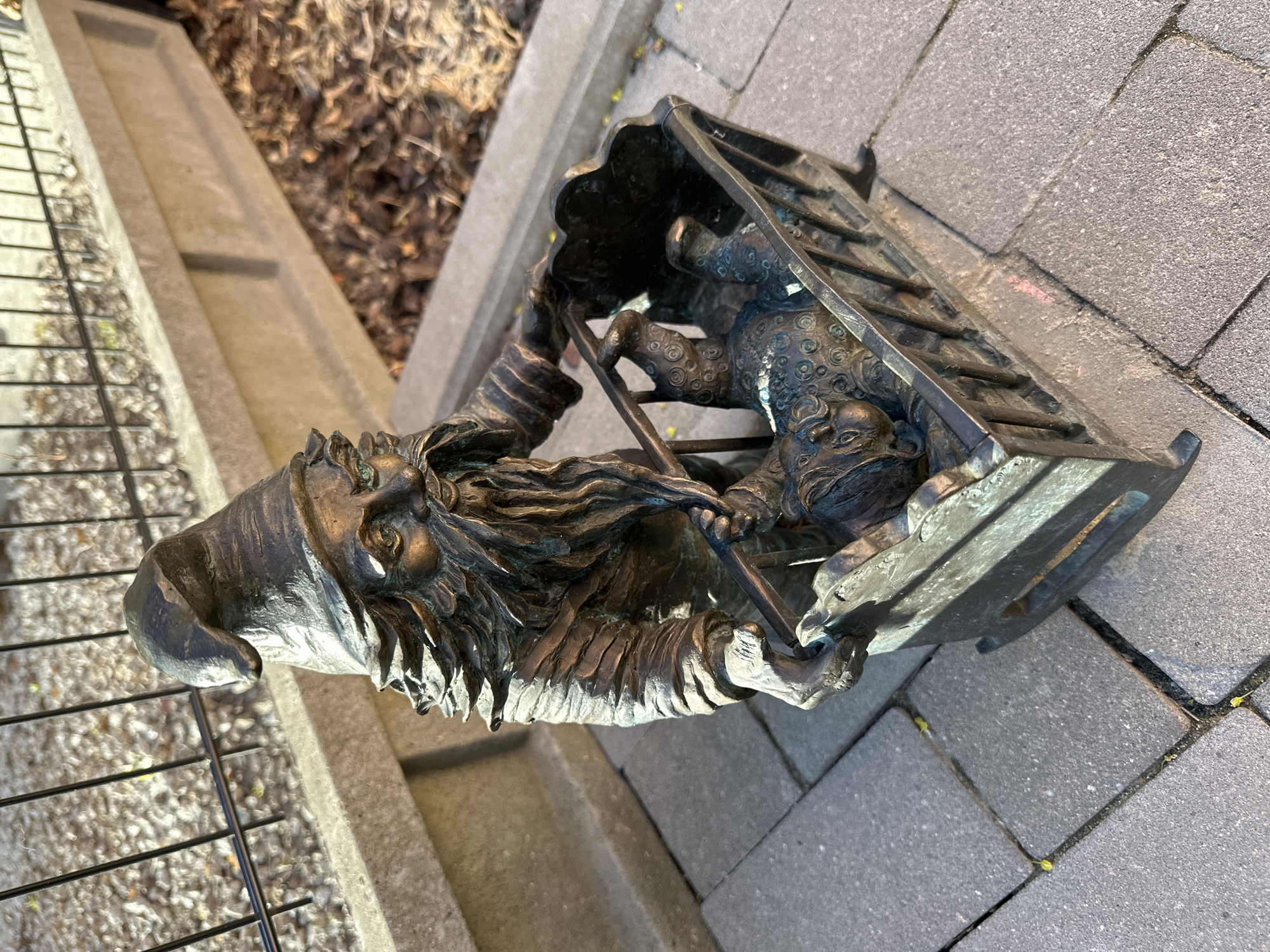 The image captures a bronze sculpture of a Wrocław dwarf rescuing a lion cub from a cage. The sculpture sits on a brick-paved area next to a concrete structure and a metal fence. Fallen leaves are visible in the background. The lion cub is lying on its back in the cage, while the dwarf is shown opening the cage door.
