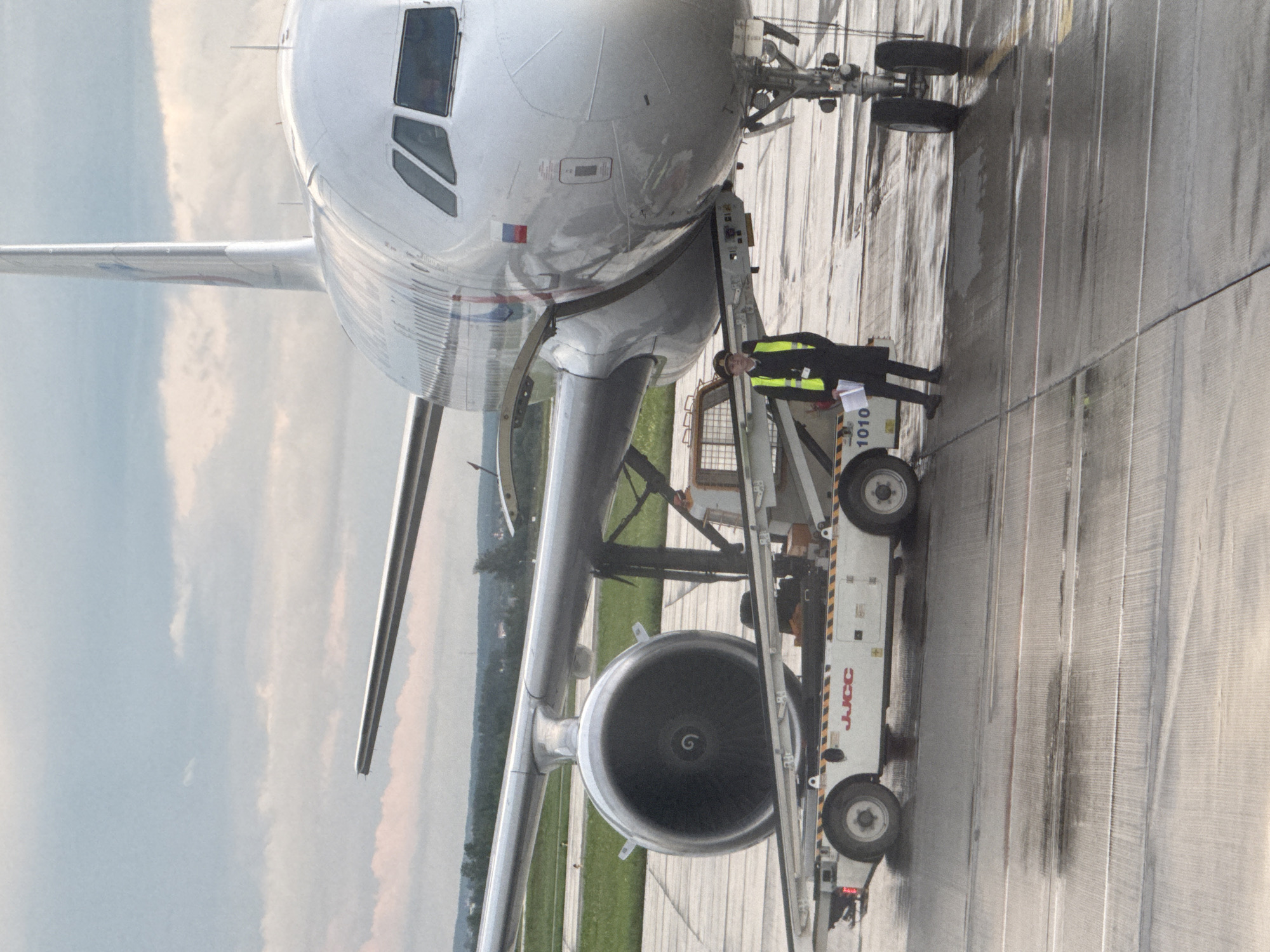 An airport ramp worker in a yellow safety vest and dark coat stands beside a pushback tractor. The tractor is attached to the front landing gear of a large, white passenger airplane. The airplane has its engines mounted under the wings. The ground appears wet, suggesting recent rain. The sky is overcast with some clouds. In the background, there is greenery and some buildings. The image was captured at Koltsovo International Airport (SVX) in Yekaterinburg, Russia.