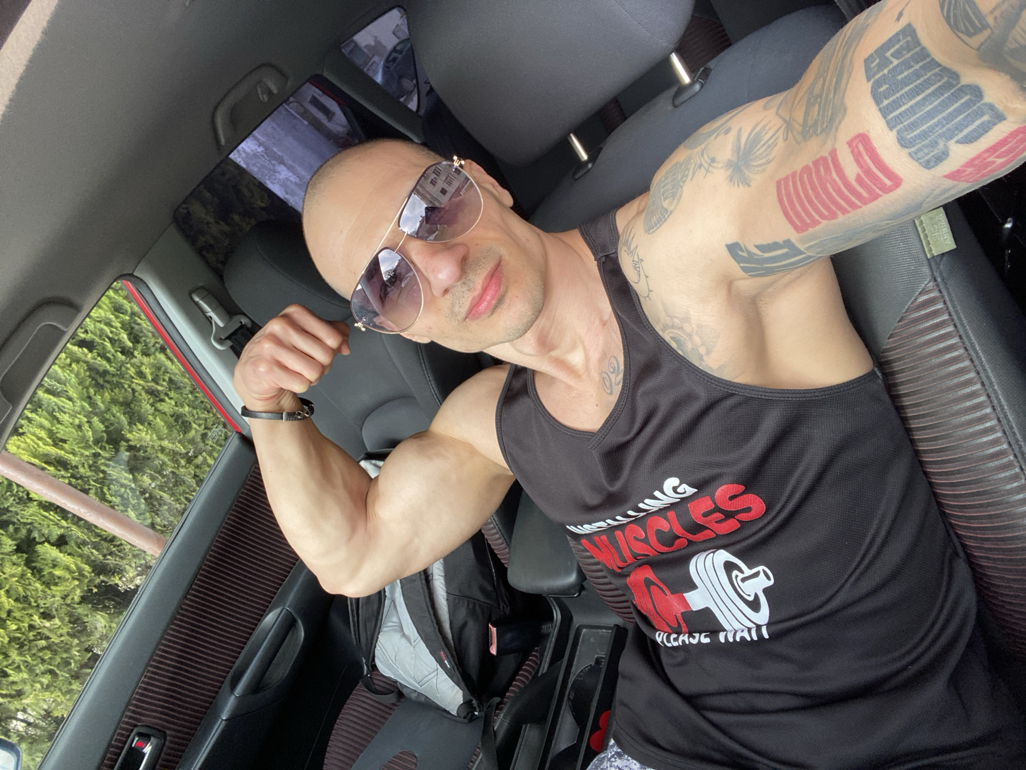 A man is posing for a selfie inside a car. He is bald, wearing sunglasses and a black tank top with a weightlifting-themed design, and flexing his bicep. He has numerous tattoos on his arms. The interior of the car includes gray leather seats, and through the window, there are green trees and a glimpse of a building. The environment appears to be outdoors, with natural light illuminating the scene. Pleven, Bulgaria is not visible in the image.
