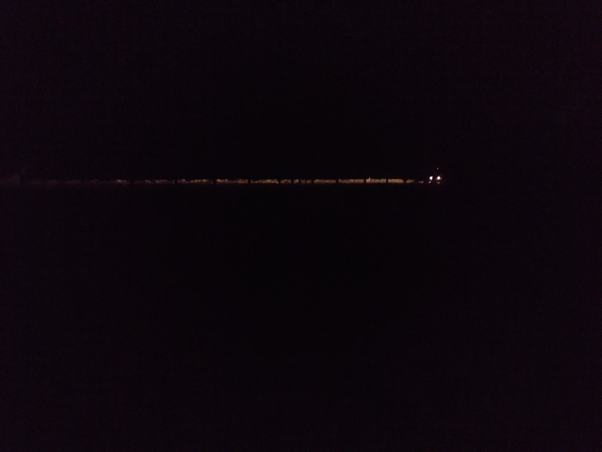 The media file presents an extremely dark, low-light image, likely captured at night. The scene is dominated by a vast expanse of black, with discernible features appearing only in a horizontal band slightly below the center. This band consists of numerous small, blurry points of yellowish-orange light, suggesting distant artificial illumination.
These lights are spread along what appears to be a distant structure or landmass, possibly a shoreline, a road, or the edge of a settlement. Towards the right side of this horizontal cluster, two brighter, more distinct white-yellow lights are visible, which could be headlights of a vehicle or powerful individual light sources. The extreme darkness of the surroundings, including the sky and foreground, indicates a nighttime setting in an area with minimal ambient light, possibly a rural or remote location like Ustyvytsia, Ukraine. No specific activities, people, or discernible text are present. The image conveys a sense of quiet vastness with a distant indication of human presence.