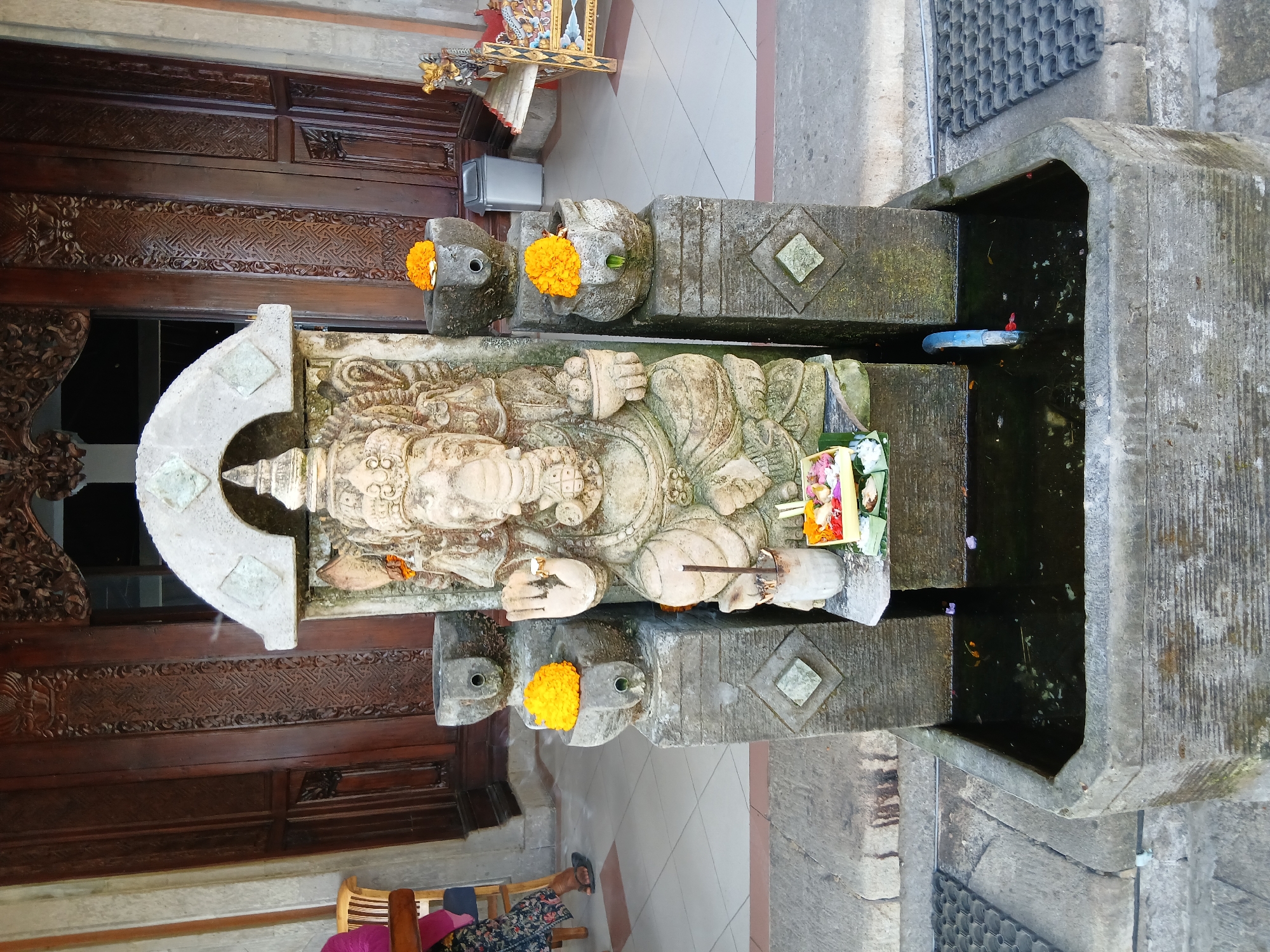 In Kintamani, Indonesia, a stone statue of Ganesha, the Hindu deity with an elephant head, is positioned in front of a large wooden door. The statue is seated and adorned with orange marigolds in two stone bowls flanking it. Below the statue, a small offering of flowers and incense sticks is placed on a tray. The statue is situated within a stone structure that extends into a water basin. The basin contains dark water with a few floating flower petals and leaves. A blue pipe extends into the water, possibly for water circulation. The environment appears to be an outdoor setting, possibly a temple or a traditional Balinese compound, with tiled flooring and architectural elements of stone and wood. In the background, a glimpse of a person sitting on a chair is visible to the left, and ornate wooden carvings are present on the door.