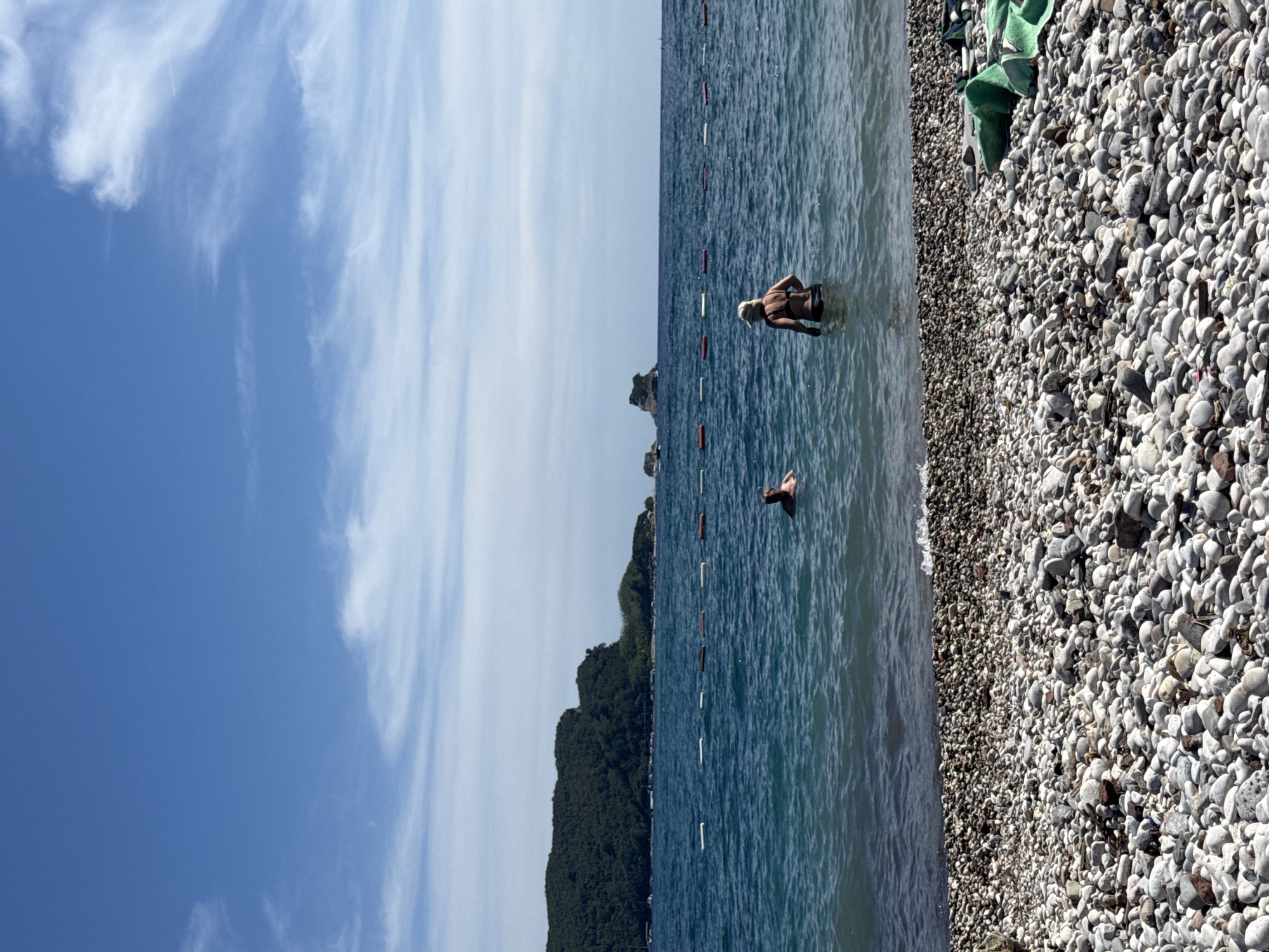 The image depicts a wide shot of a rocky beach leading into the Adriatic Sea. The sky is a clear blue with wispy white clouds. In the foreground, a pebble-strewn beach is visible with some green fabric objects on the right. The sea is a vibrant blue, with a line of orange and white buoys marking an area in the water. Two individuals are present in the sea; one person wearing a hat and a dark swimsuit is standing in the water facing away from the camera, while another person is swimming further out. In the background, a densely forested green hill forms a prominent landmass, and several rocky islets are scattered in the distance. The overall scene suggests a sunny day at the beach in a coastal area, likely near Budva, Montenegro, given the characteristic rocky shores and distant islets.