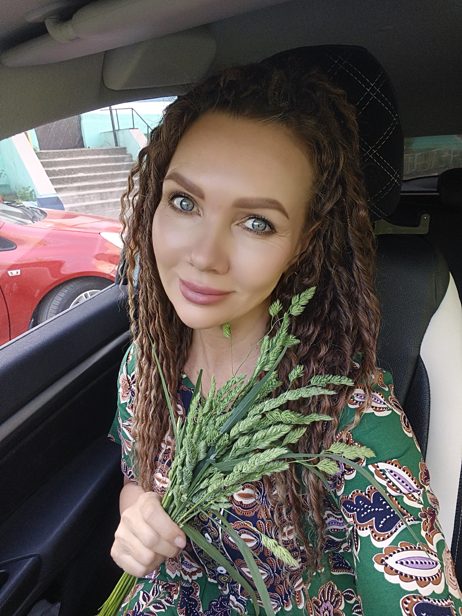 A medium shot shows a woman inside a car holding a bunch of green grass. The woman has curly brown hair and is wearing a green dress with a paisley-like pattern. Her skin is fair, and she has light blue eyes and pink lipstick. The background through the car window shows a red car, concrete stairs, and a building painted in a light blue color. The car's interior is visible, with a black headrest and a white panel on the seat.