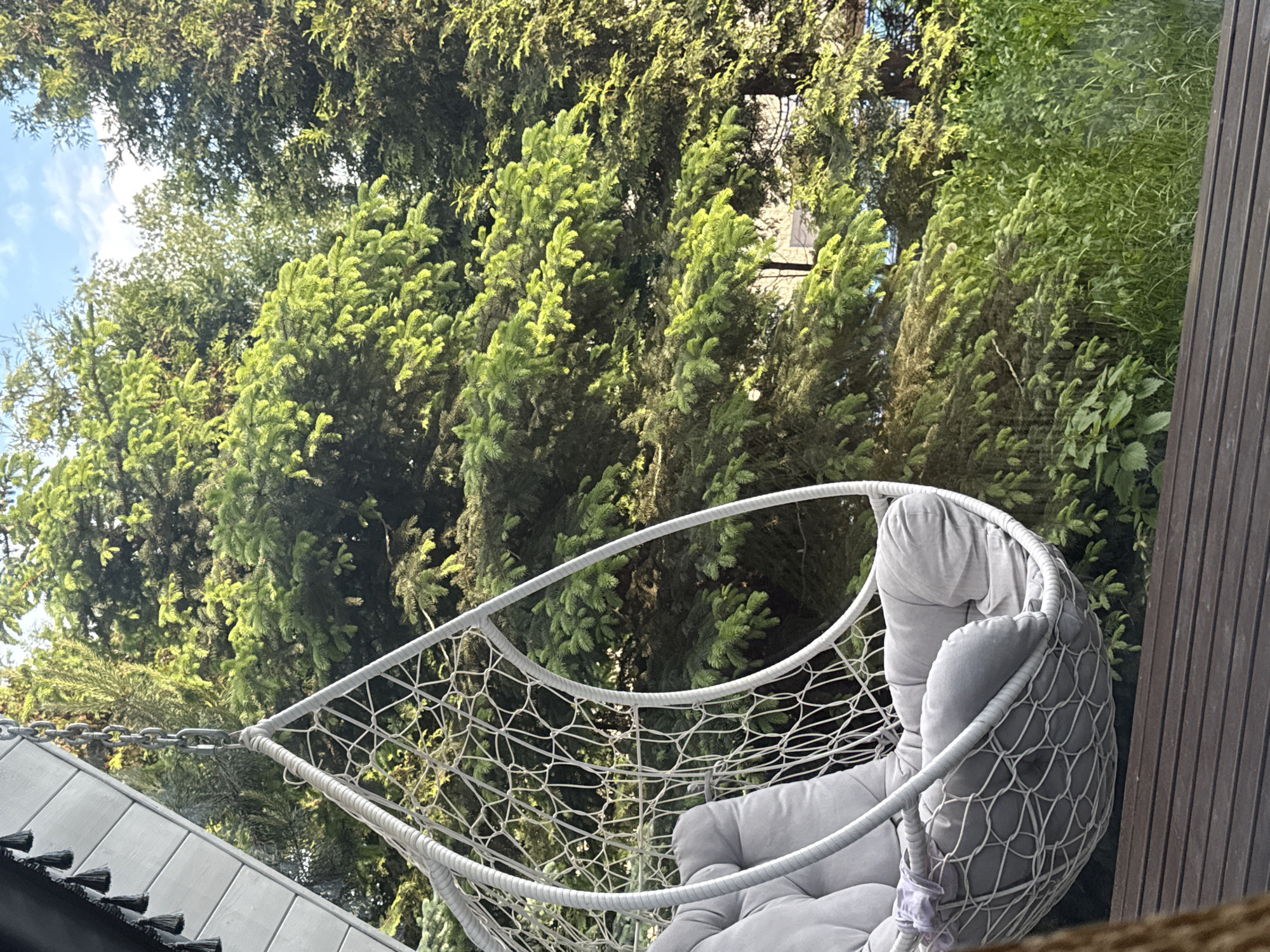 The image depicts a hanging chair situated outdoors, possibly a patio or backyard, with lush green trees forming the backdrop. The chair is white with a net-like pattern and soft grey cushions. Adjacent to the chair is a dark wooden structure, likely a wall or fence. A glimpse of blue sky is visible through the trees. The location is specified as Чёрное, Russia.