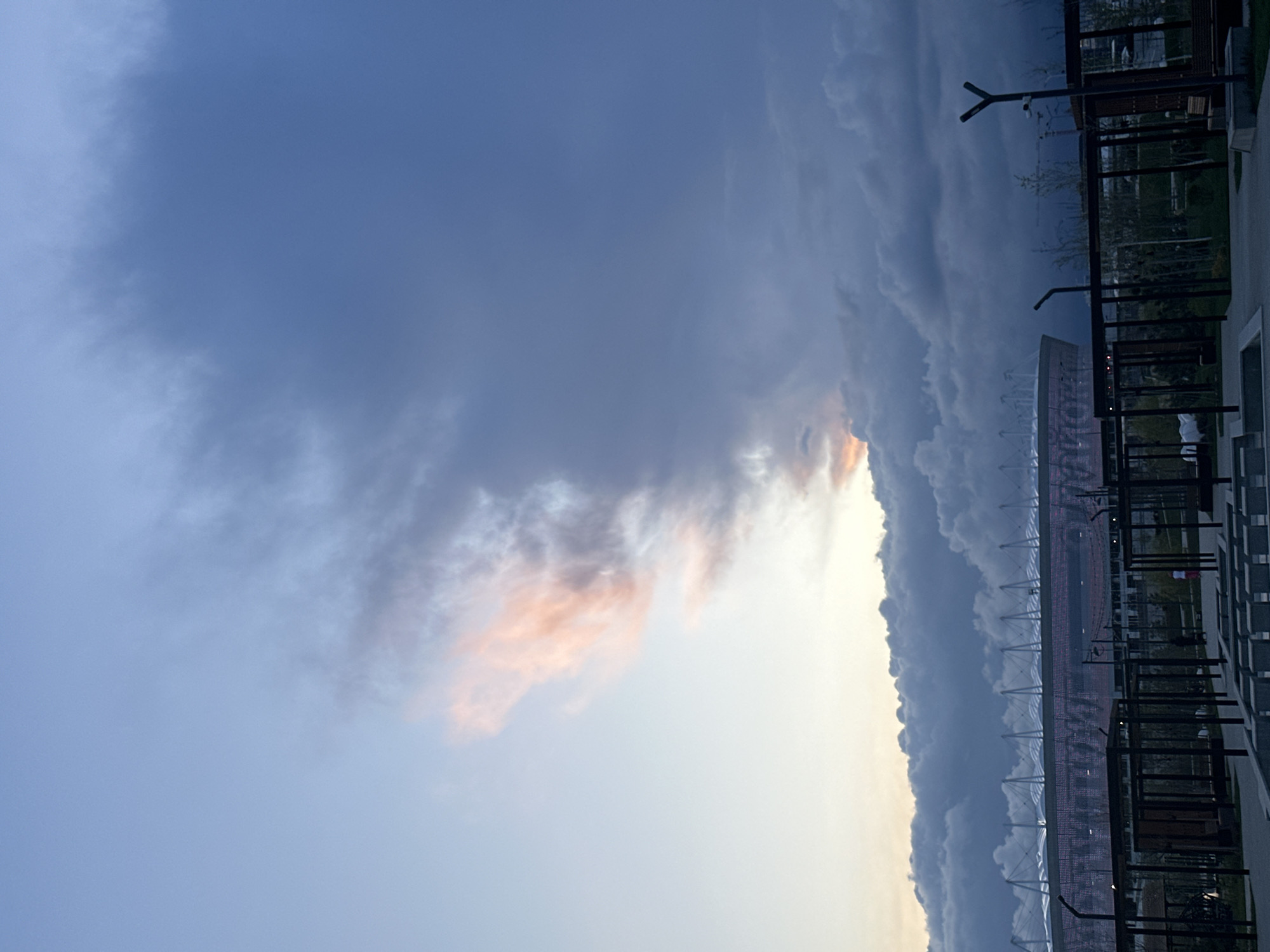 The image captures a view in Rostov-on-Don, Russia, under a sky dominated by dramatic clouds. The sky transitions from a light, clear expanse to heavy, dark clouds, with some clouds tinged with a peach color. A stadium, identifiable by its architectural structure of repeating arches, stands out against the sky. A metal structure with vertical and horizontal lines, possibly part of a building or an outdoor structure, is visible in the foreground.