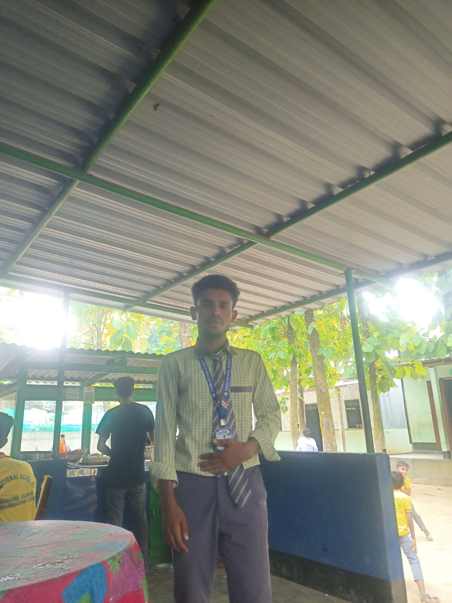 The image shows a young man standing under a corrugated metal roof supported by green metal beams. He is wearing a checkered shirt, grey pants, and a tie with a lanyard containing an identification card. The man is looking towards the camera. In the background, there are trees and several people, some of whom are blurred or partially obscured. One person is wearing a yellow shirt with text that reads "VISION NATIONAL ACADEMY". Another person in the background appears to be working at a counter. The location is described as Hatsinigmari, India, suggesting an outdoor or semi-outdoor area, possibly a school or public space with a covered seating area. The lighting suggests it is daytime.
