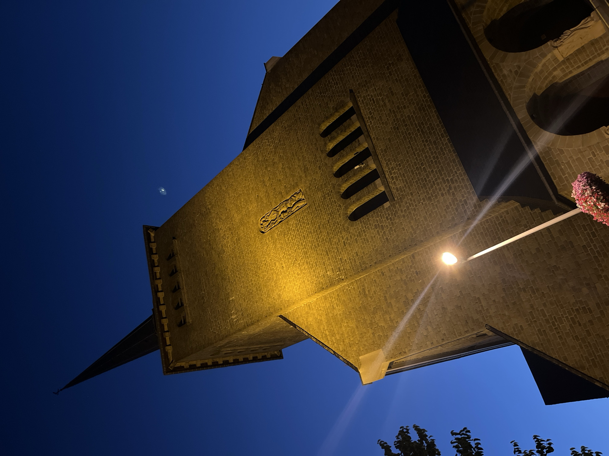 The image shows a close-up, low-angle view of the brick bell tower of the Saint Vith Basilica in Saint-Vith, Belgium, at dusk. The tower is illuminated by a street lamp, casting a warm yellow glow on the brickwork. The sky is a deep blue, with a faint crescent moon visible in the upper right quadrant of the image. The spire of the tower is dark and pointed, reaching towards the sky. Decorative elements, such as arched windows and a carved relief, are visible on the tower's facade. A street lamp with a bright light is positioned in the foreground, with light rays emanating from it. To the left of the tower, a tree with dark leaves is partially visible. The bottom right of the image shows arched entrances to the basilica. A cluster of pink flowers is visible at the base of the street lamp. The scene depicts the exterior of a religious building illuminated at twilight.