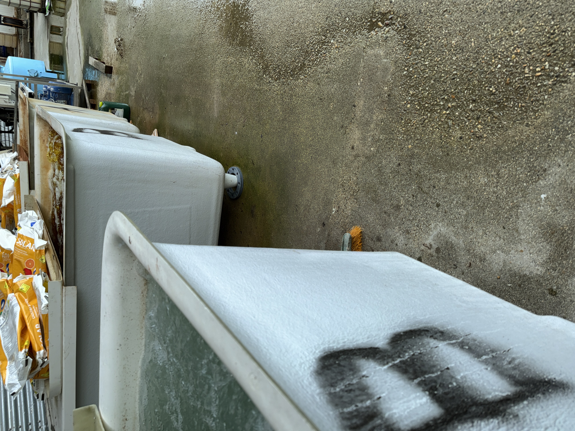 The image shows an outdoor area in what appears to be an industrial or storage yard. The dominant features are a series of white, rectangular containers or tanks lined up. Some have black markings or dirt on the top surfaces. One of the tanks has a grey metal outlet pipe attached. To the left of the tanks is a stack of cardboard boxes, likely containing items labeled 'SOLAR'. The ground is paved and appears to be wet, showing a textured surface with embedded gravel. In the background, there are glimpses of other structures and items, including a blue container. The overall environment suggests an outdoor storage or working area. The presence of Cyrillic on the packaging suggests the scene is in Bulgaria, such as in the city of Stara Zagora.