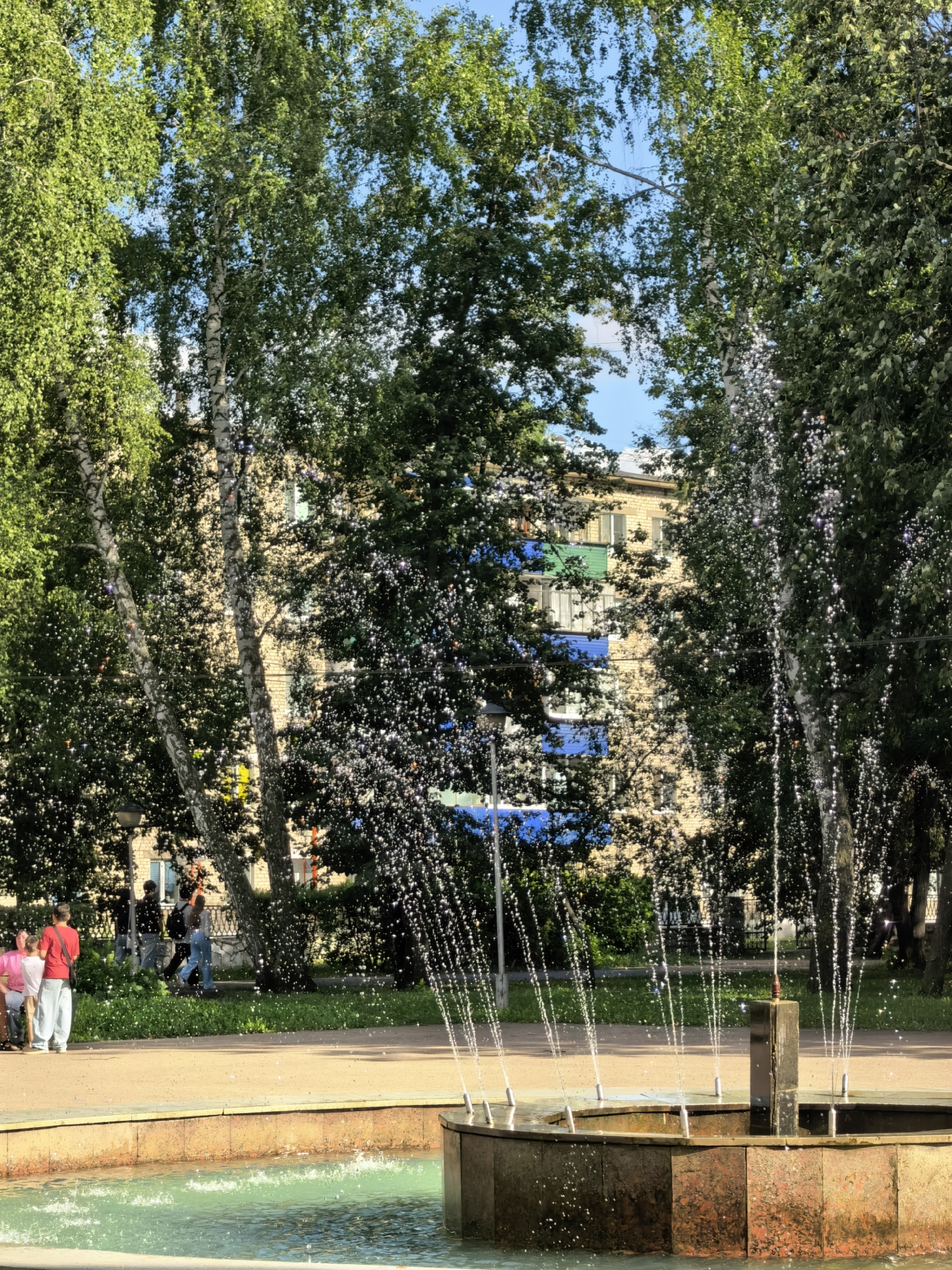 The image depicts a sunny day in a park in Sterlitamak, Russia. In the foreground, a stone fountain with turquoise water is active, with streams of water spraying upwards and catching the sunlight. Beyond the fountain, a paved pathway runs horizontally, separating the park's grassy area from the fountain. Several people are visible on the pathway and in the grassy area. To the left, three individuals, two women and one man, are standing near a bench. Further to the right, a group of four people, including two men and two women, are walking away from the viewer. In the background, tall trees with green foliage create a natural screen, with a multi-story building partially visible behind them. The building has a light-colored facade with bands of blue and green paint. The sky is a clear blue.