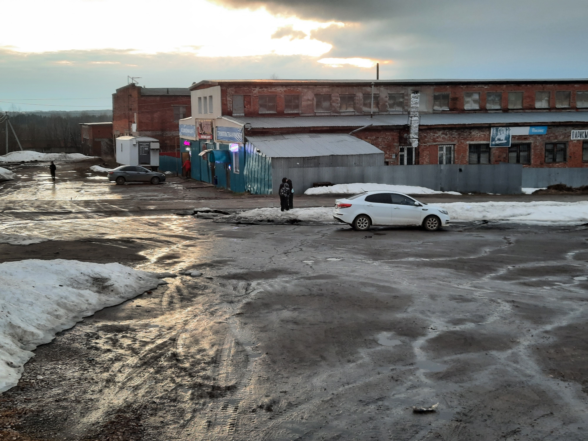 The image depicts a thawing scene in Krasnogorskoye, Russia. Patches of snow remain on the ground, mixed with muddy areas and puddles. A white car is parked on the right side of the scene. A grey car is parked left of frame. Two people are standing in front of a building with a covered market or shop, with red and blue canopies. Another person is standing further left. The buildings are a mix of brick and corrugated metal. The sky is overcast. The environment is urban, with signs of infrastructure.