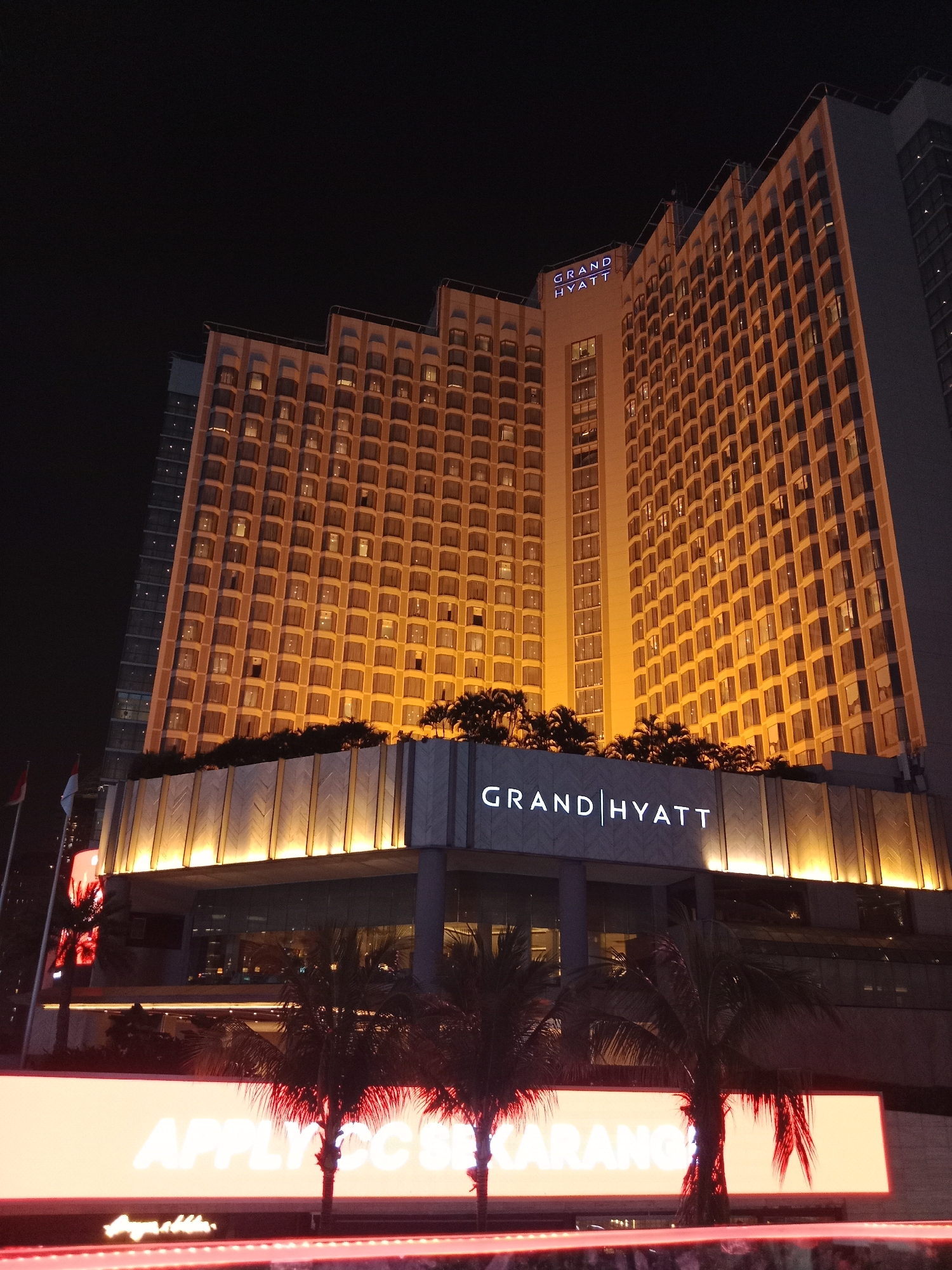 An exterior, eye-level, full-shot photograph taken at night depicts the Grand Hyatt hotel in Central Jakarta, Indonesia. The brightly lit hotel is tall with many windows that are illuminated from within. Palm trees are in the foreground with a bright orange sign that reads "APPLIC SC KARANG" in large white letters. The Grand Hyatt logo is visible on the hotel building. There are flags on the left side of the image.