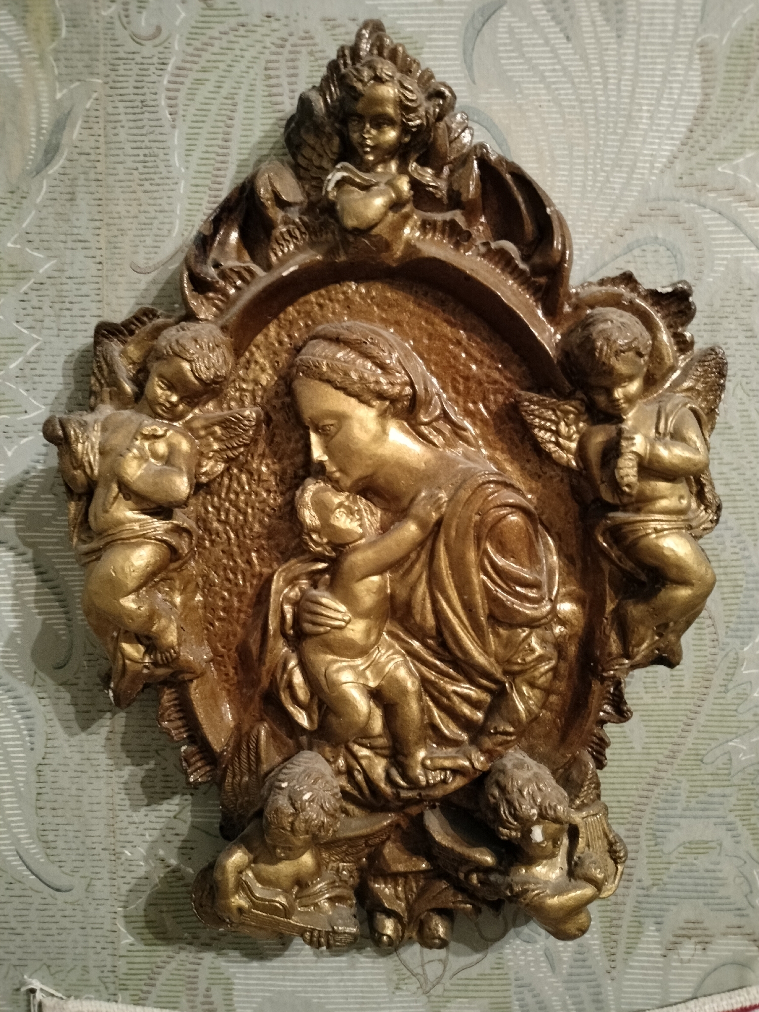 An antique, gilded relief depicts the Madonna and Child embraced, surrounded by cherubs. The central figures are rendered with soft, flowing lines, with the Virgin Mary gazing tenderly at the infant Jesus. Several cherubic figures, with wings outstretched, are positioned around them, some playing musical instruments or holding symbolic objects. The artwork is set within an ornate, decorative frame that echoes the style of the relief. The background is a textured wallpaper with a subtle, pale green floral pattern. The image is a close-up, vertical shot of this devotional piece, likely a wall hanging, emphasizing its intricate detail and aged patina. The object is located on a wall in a room in the city of Lykhove, Ukraine.