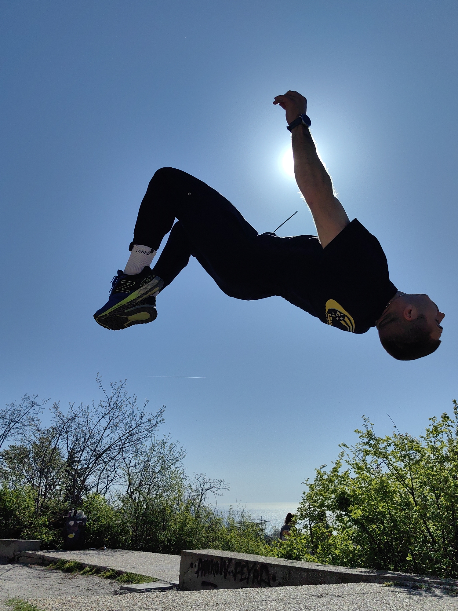 The image shows a person in mid-air, performing a backflip against a bright blue sky with the sun visible. The person is wearing a black t-shirt, black pants, and running shoes with white socks that have the word "LOSER" printed on them. The person is wearing a watch on their left wrist.
The background includes trees and greenery at the bottom of the frame. A concrete platform is visible in the foreground, and the ocean is visible in the background between the trees. A fire hydrant is visible between the trees on the left of the frame. There is graffiti on the concrete platform in the foreground.