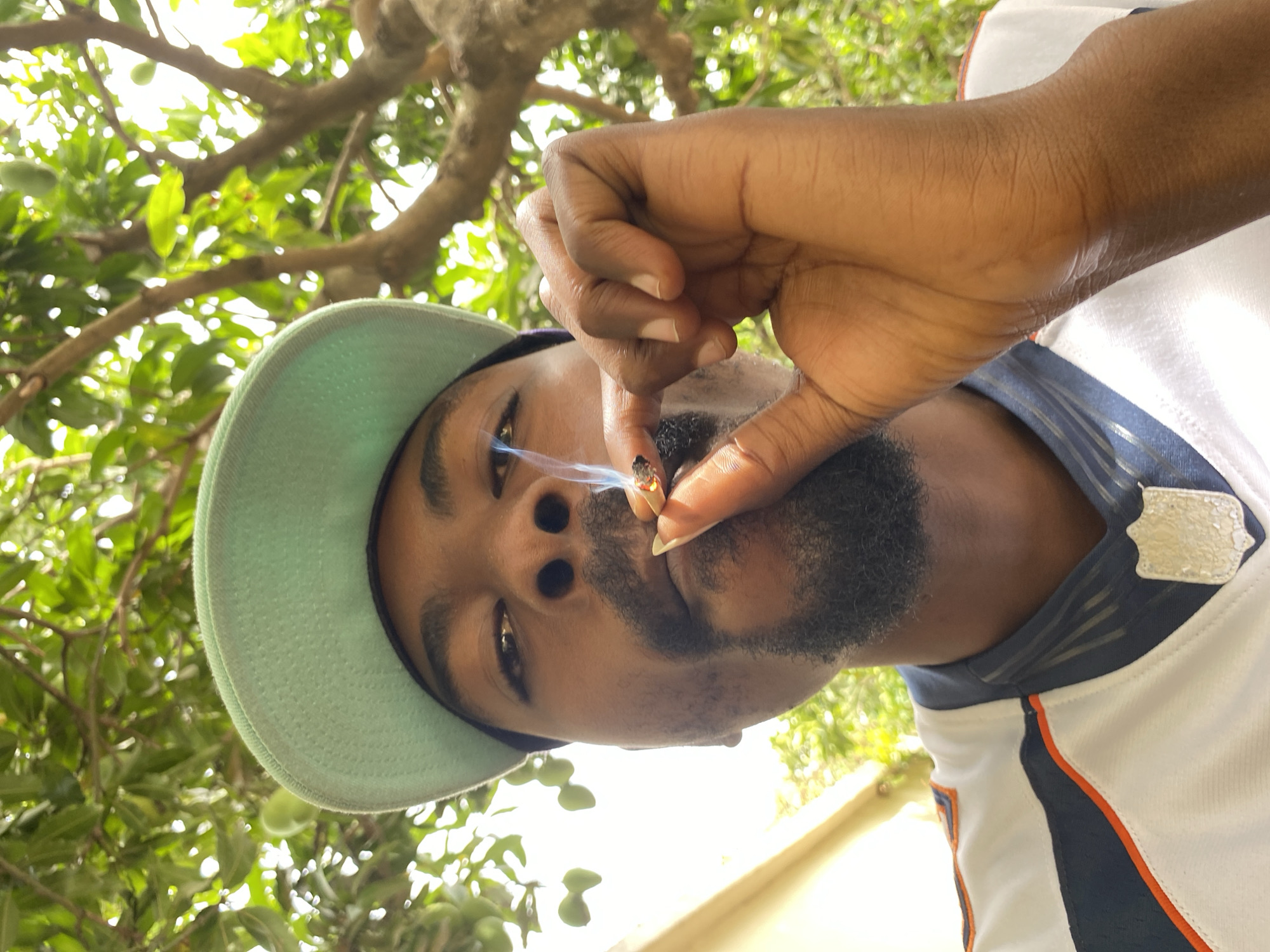 The image shows a Black male with a goatee and mustache, wearing a light green baseball cap and a white sports jersey with navy and orange trim, holding a lit object between his fingers that is emitting smoke. He is outdoors under a tree with green foliage. The location is presumed to be Jos, Nigeria.